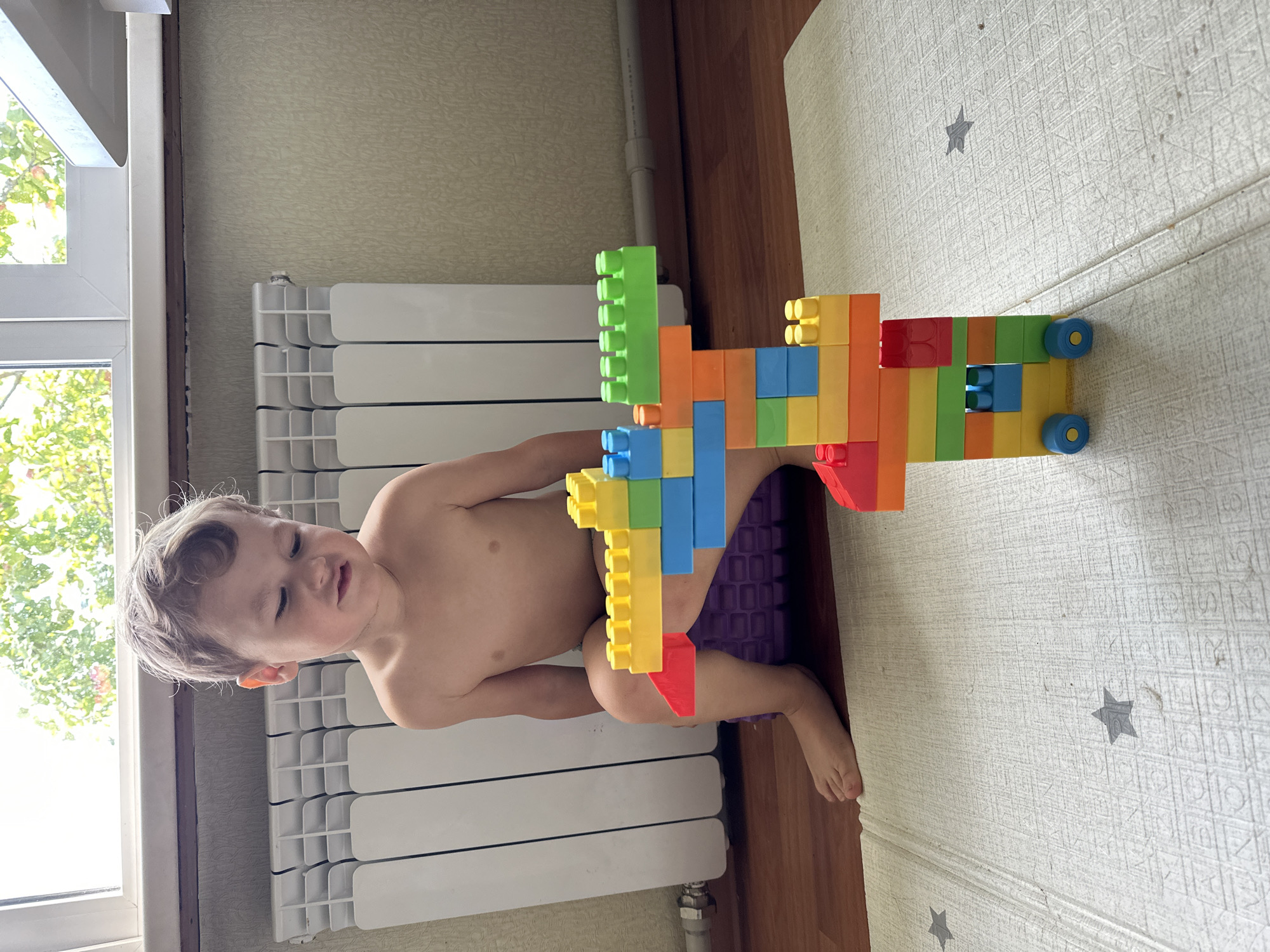 A young boy with blonde hair, wearing no shirt, sits on a purple mat with his legs bent. He is looking down at a colorful toy structure made of building blocks, which appears to be a giraffe. The blocks are red, yellow, orange, green, and blue. The boy's facial expression suggests he is focused or possibly making a funny face.
The background shows a white radiator with vertical fins, a light-colored wall with a textured wallpaper, and a window with sunlight streaming through, illuminating green foliage outside. The floor is wooden, and a light-colored play mat with a grid pattern and some star shapes is visible in the foreground. The image appears to be taken indoors, likely in a residential setting.
The image does not contain enough information to determine its location in Nizhny Novgorod, Russia.