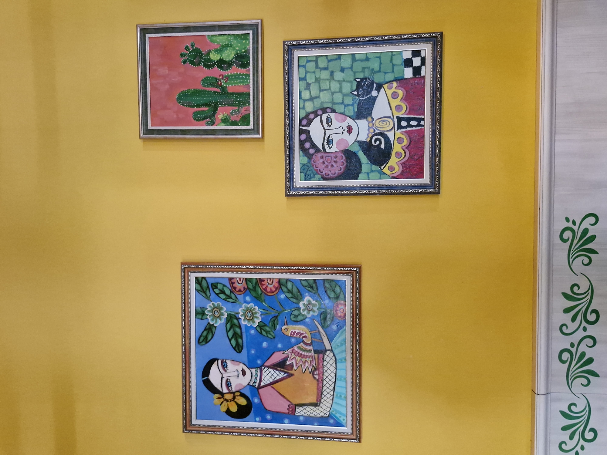 The image shows a wall painted a bright yellow color. Three framed paintings hang on the wall. The painting on the top left depicts a cactus against a red background. The painting on the top right features a stylized portrait of a woman with a black cat on her head, set against a background of green and checkered patterns. The bottom painting shows another stylized portrait of a woman with flowers and a bird. A decorative white molding with green floral designs is visible on the right edge of the image.