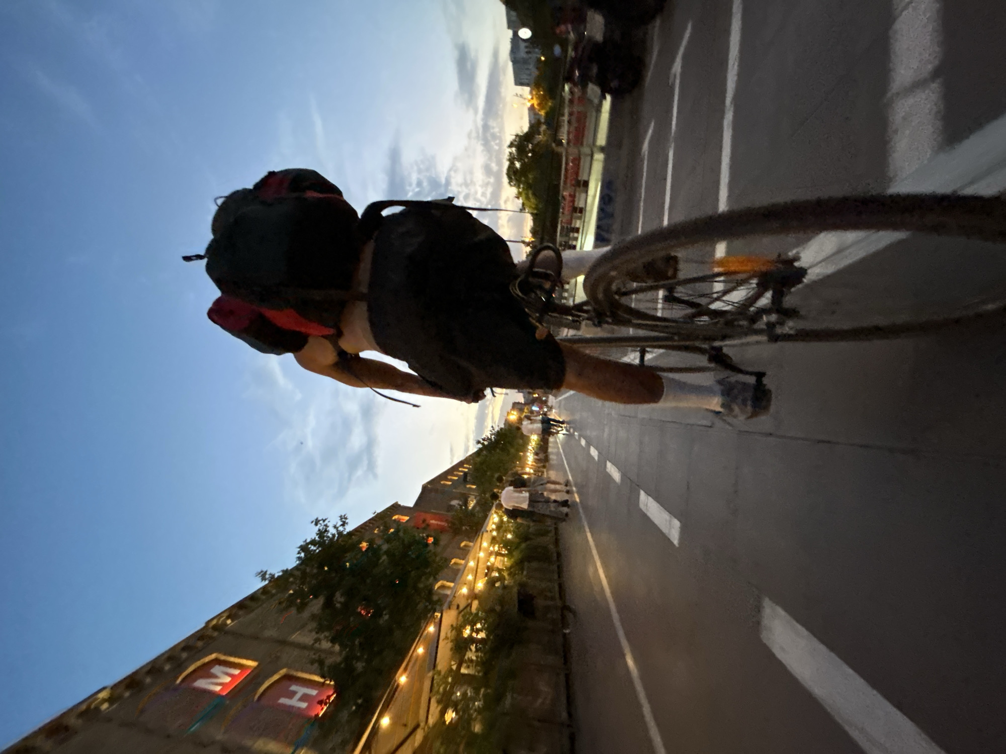 The image shows a low-angle, wide-shot of a person riding a bicycle on a street at dusk in Barcelona, Spain. The cyclist is seen from behind, wearing a backpack and black shorts. The street is paved and marked with white lines, suggesting a bike lane. On the left side of the street, there's a building with illuminated signs displaying the letters "M" and "H". Adjacent to the building, there are outdoor seating areas with string lights, indicating a commercial establishment like a restaurant or bar. Other pedestrians can be seen further down the street, also walking or cycling. In the background, there are more buildings and some trees, with a bridge-like structure visible in the far distance. The sky is a deep blue with some lighter clouds, characteristic of twilight. The overall impression is of a vibrant urban environment during a transition from day to night.