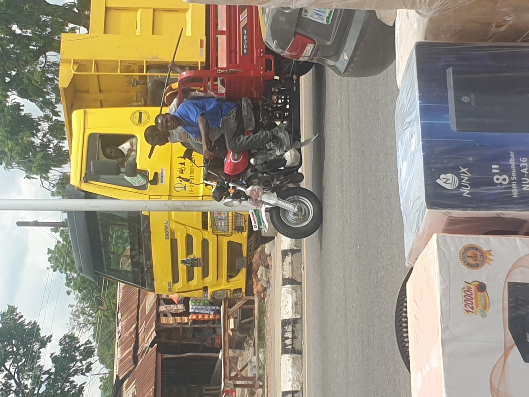 The image depicts an outdoor scene in an urban setting, likely a city in an unknown location. Two people are riding a white motorcycle on a road. One is wearing a blue shirt, and the other a light grey shirt. A large yellow truck is stopped nearby, with a man visible in the driver's seat. A red truck is also visible, partially behind the motorcycle. The background includes buildings with corrugated iron roofs, trees, and utility poles. In the foreground, there are boxes of electronics, including a "NUNIX" soundbar, suggesting a shop or storage area. The scene suggests a busy street with various vehicles and commercial activity.