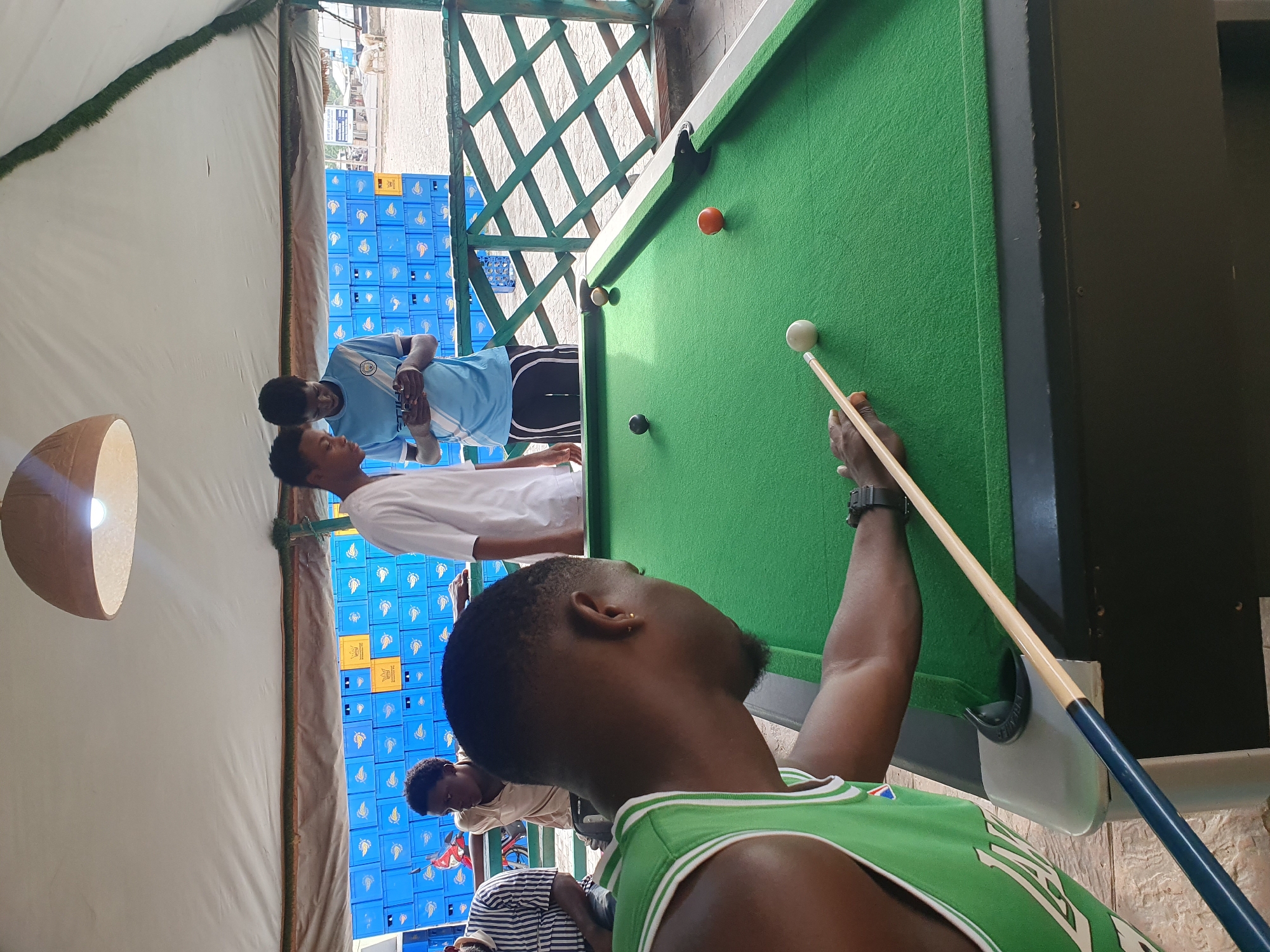 A group of people is playing pool in an outdoor setting in Lokoja, Nigeria. The pool table is green and has several pool balls on it. One man is in the foreground, holding a pool cue and aiming at the cue ball. He is wearing a green and white tank top and a wristwatch. Behind him, other people are watching the game. A stack of blue plastic crates is visible in the background. The setting appears to be semi-enclosed, possibly a covered patio or outdoor bar area.