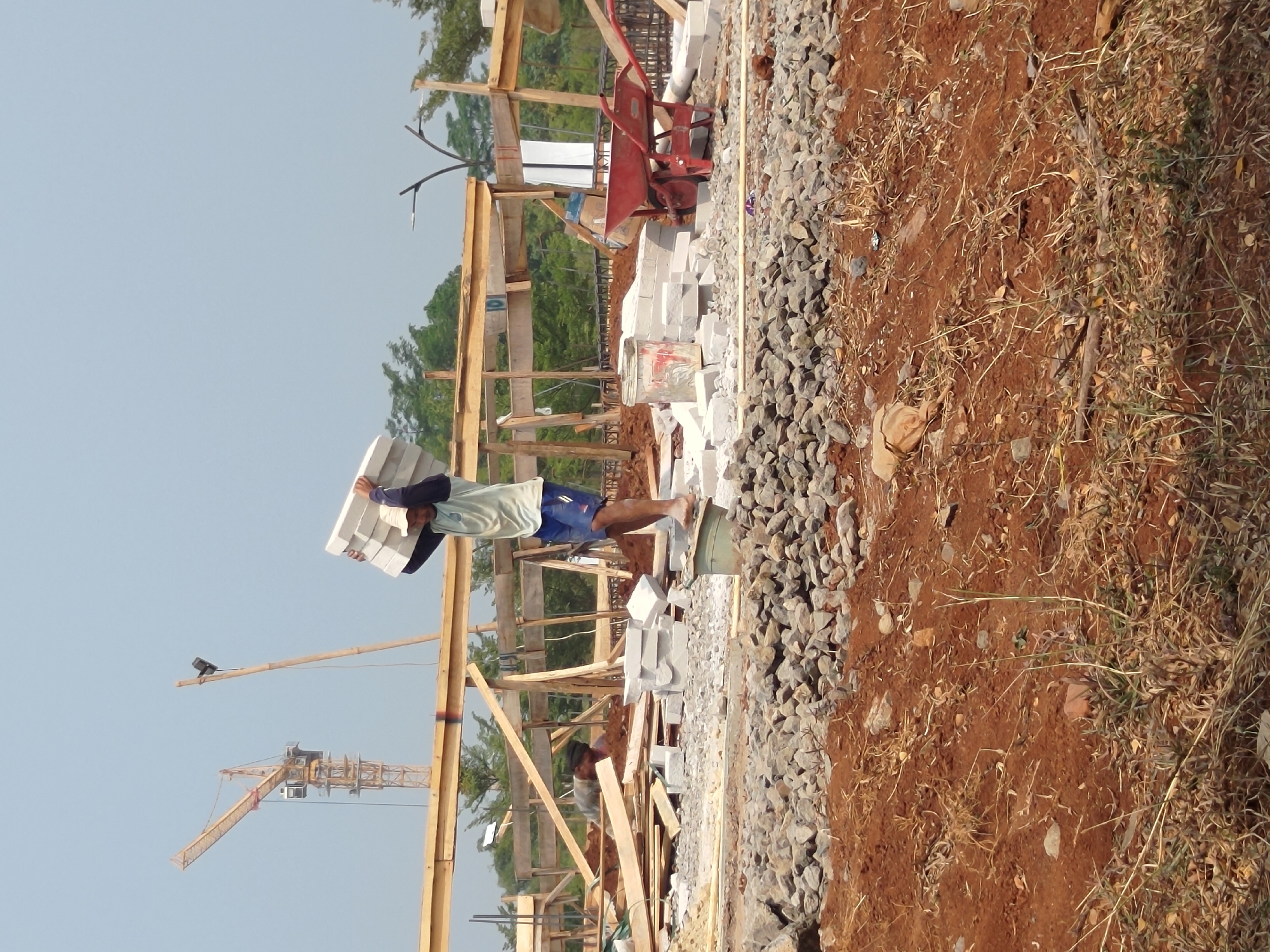 The image depicts a construction site in Depok, Indonesia, on a bright, overcast day. In the foreground, reddish-brown soil is covered with scattered rocks and dry vegetation. In the mid-ground, a man is carrying a stack of rectangular blocks on his head, wearing a light blue t-shirt and dark blue shorts. He is barefoot and stands on a green bucket. Behind him, the construction site is in progress with wooden scaffolding and piles of white blocks. A red wheelbarrow is visible to the right of the blocks. In the background, a large yellow tower crane is present, along with a clear sky. The overall scene captures a moment of manual labor at a construction project under development.