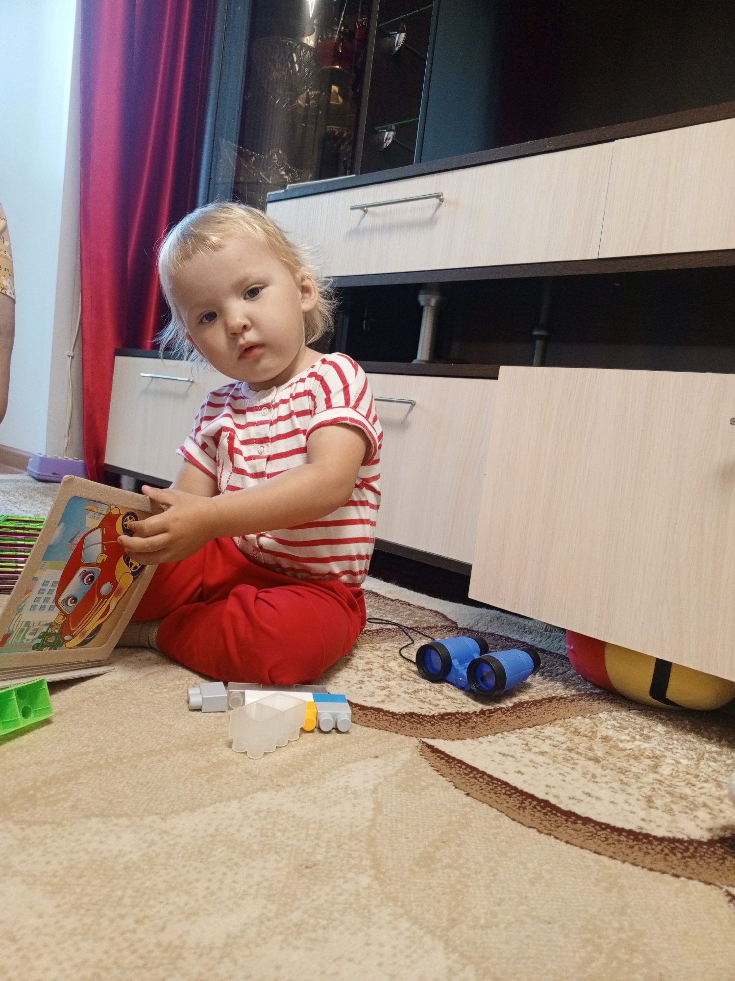A young Caucasian child with blonde hair, wearing a red and white striped shirt and red pants, sits on a beige carpet. The child is holding a wooden puzzle with a cartoon car on it. In front of the child are some building blocks and a pair of blue toy binoculars. Behind the child is a wooden cabinet. A red curtain is visible on the left side of the image. The scene appears to be indoors, possibly in a home. The image is taken from a low angle, looking up at the child. The location is not specified in the image or the provided context, so it cannot be determined if it is in Dyurtyuli, Russia.