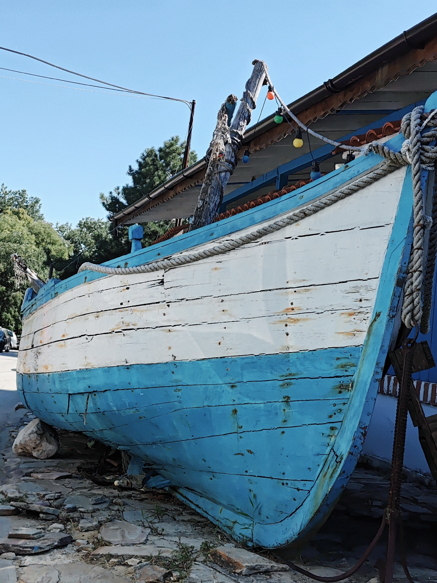 A weathered, blue and white wooden boat is propped on chocks on a gravelly surface. The boat is angled upwards, its bow facing the viewer. The hull is constructed of horizontal wooden planks, some of which are cracked and show signs of wear. A thick rope is tied to the bow and draped along the top edge of the boat. Above the boat, a wooden roof with red tiles extends from a building. Colorful decorative lights are strung across the roof. Trees are visible in the background, and a blue sky fills the upper portion of the image. A car is partially visible on the left side of the frame. The scene suggests a seaside or coastal location, possibly a port or a decorative display.