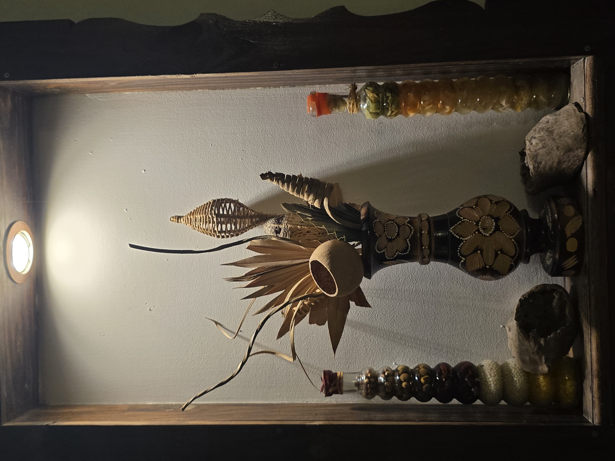 A wooden shelf, possibly mounted on a wall in Berkovitsa, Bulgaria, contains a collection of decorative items. The shelf's structure has a light shining from the left, illuminating a beige-painted wall. The objects within the shelf consist of arrangements of dried plant materials, including seed pods, dried leaves, and branches. Two glass tubes, each filled with assorted seeds or beans, are placed near the top and bottom edges of the shelf. A decorated wooden spindle or baluster, featuring painted floral designs, is centrally positioned. A rock is positioned on the right near the back. The shelf appears to be a decorative display, likely handmade or assembled from various natural and crafted elements.