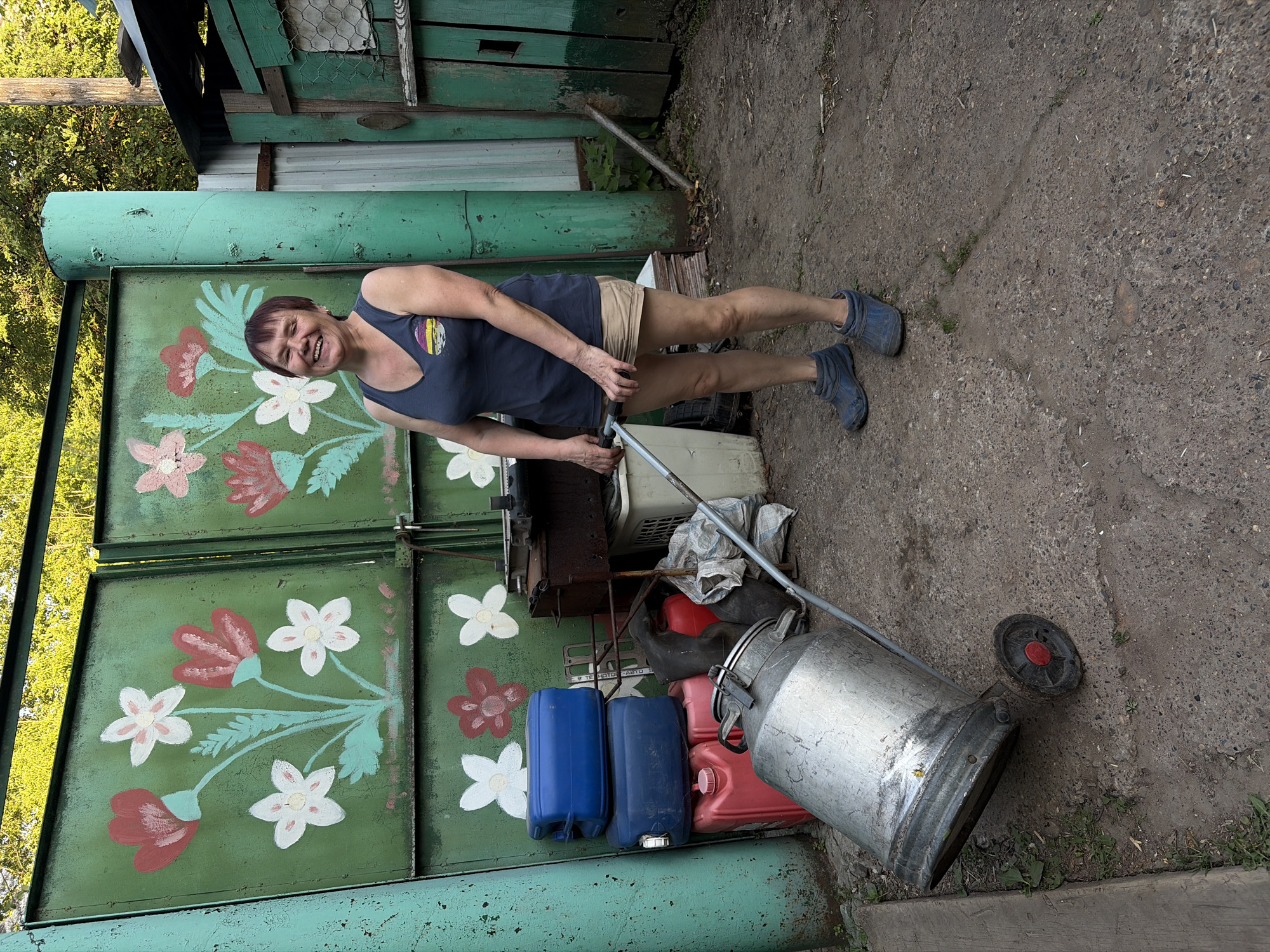 A middle-aged woman stands in a rural setting near a painted gate in what appears to be Izhevsk, Russia. The woman is wearing a dark tank top, khaki shorts, and blue work boots. She has short, dark hair and is smiling. The gate is green and painted with white and red flowers. Next to the gate are various objects, including blue and red plastic containers, a metal milk can, and a gray wheel. The ground is made of cracked concrete. The background includes trees and a wooden structure. The scene is somewhat cluttered and appears to be a working area or yard.