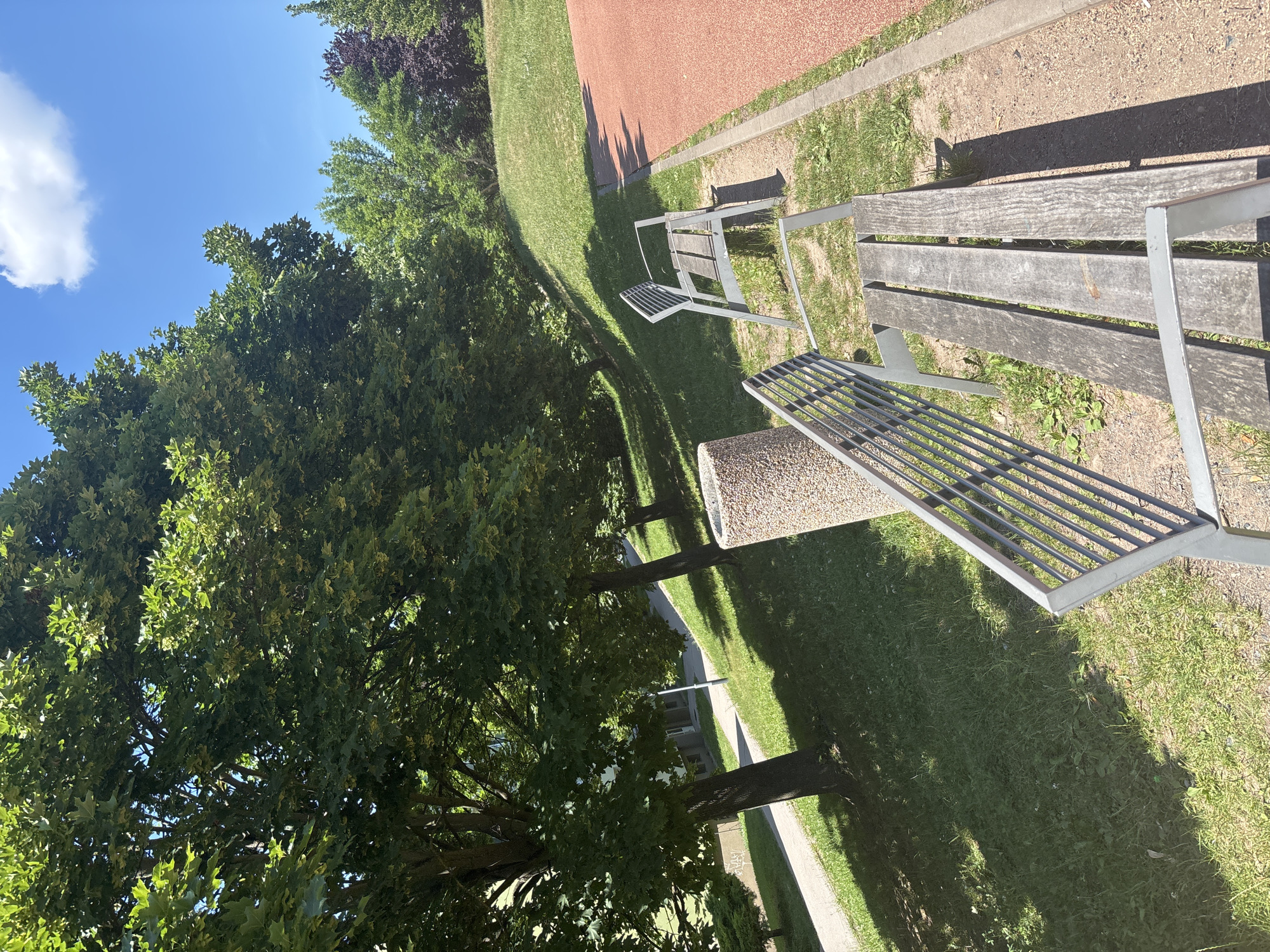 The image depicts an outdoor scene in a park-like setting, possibly a recreational area. In the foreground, two park benches made of weathered wood and metal frames are visible. The bench on the right is closer to the camera, showing its slatted wooden seat. The bench on the left is slightly further away and angled. Behind the benches, a pathway winds uphill, bordered by a grassy slope. Several mature trees with lush green foliage line the pathway, casting shadows on the grass. A cylindrical concrete trash receptacle is positioned near the pathway between the two benches. Further in the background, more trees and a red athletic track are visible, suggesting a park or sports facility. The sky above is blue with a few white clouds. The scene appears to be bathed in bright sunlight. There are no people present in the image.