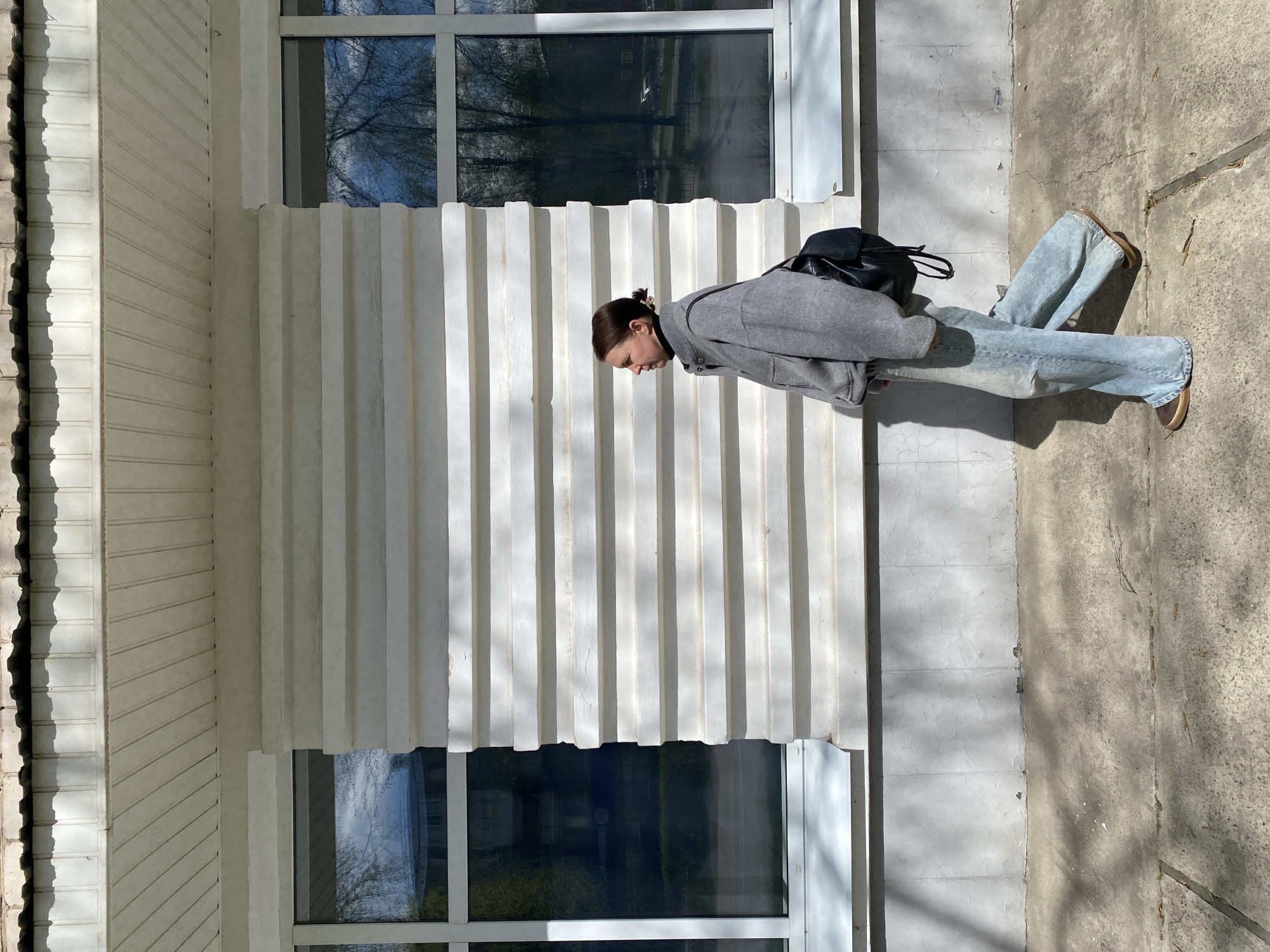 A woman stands on a concrete sidewalk in front of a light-colored building with a modern facade. She is wearing a gray jacket, light blue jeans, and brown shoes. A black backpack is slung over her shoulder. The building has a distinctive design with vertically ridged columns above and below a pair of windows, which reflect trees. Based on the architecture and surroundings, the location is likely in the city of Chaikovsky, Russia.