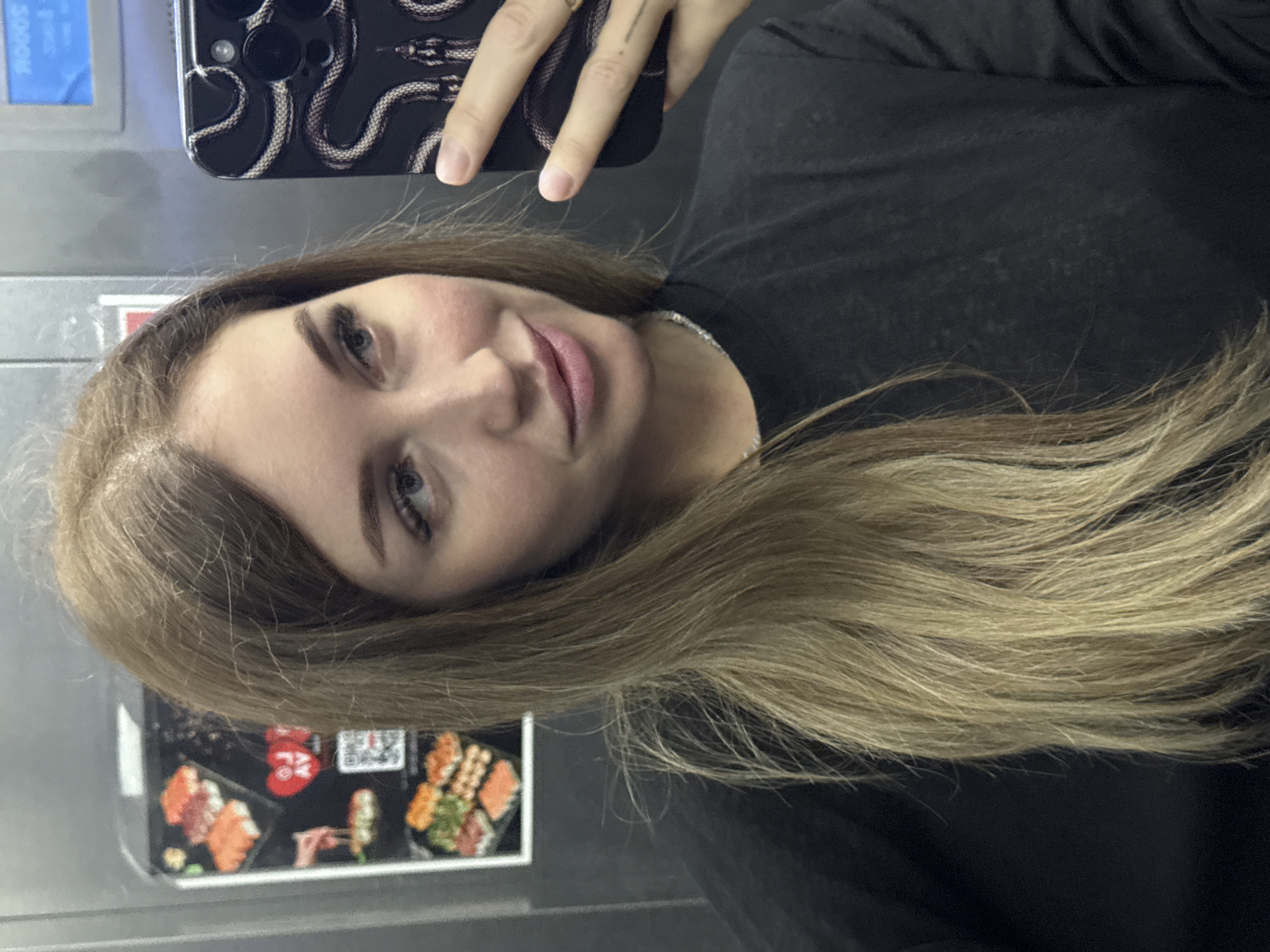 A medium shot shows a young woman with blonde hair taking a selfie in an elevator. She is wearing a black t-shirt. In the background, there are two posters, one featuring sushi and the other featuring a snake pattern. The elevator doors are metallic gray. The floor of the elevator is not visible. There is a sign on the wall that is partially obscured, but appears to be digital. The woman is holding a smartphone with a snake-patterned case. The image is a mirror selfie, and the woman is looking at the camera. The image was taken in Chaikovsky, Russia.