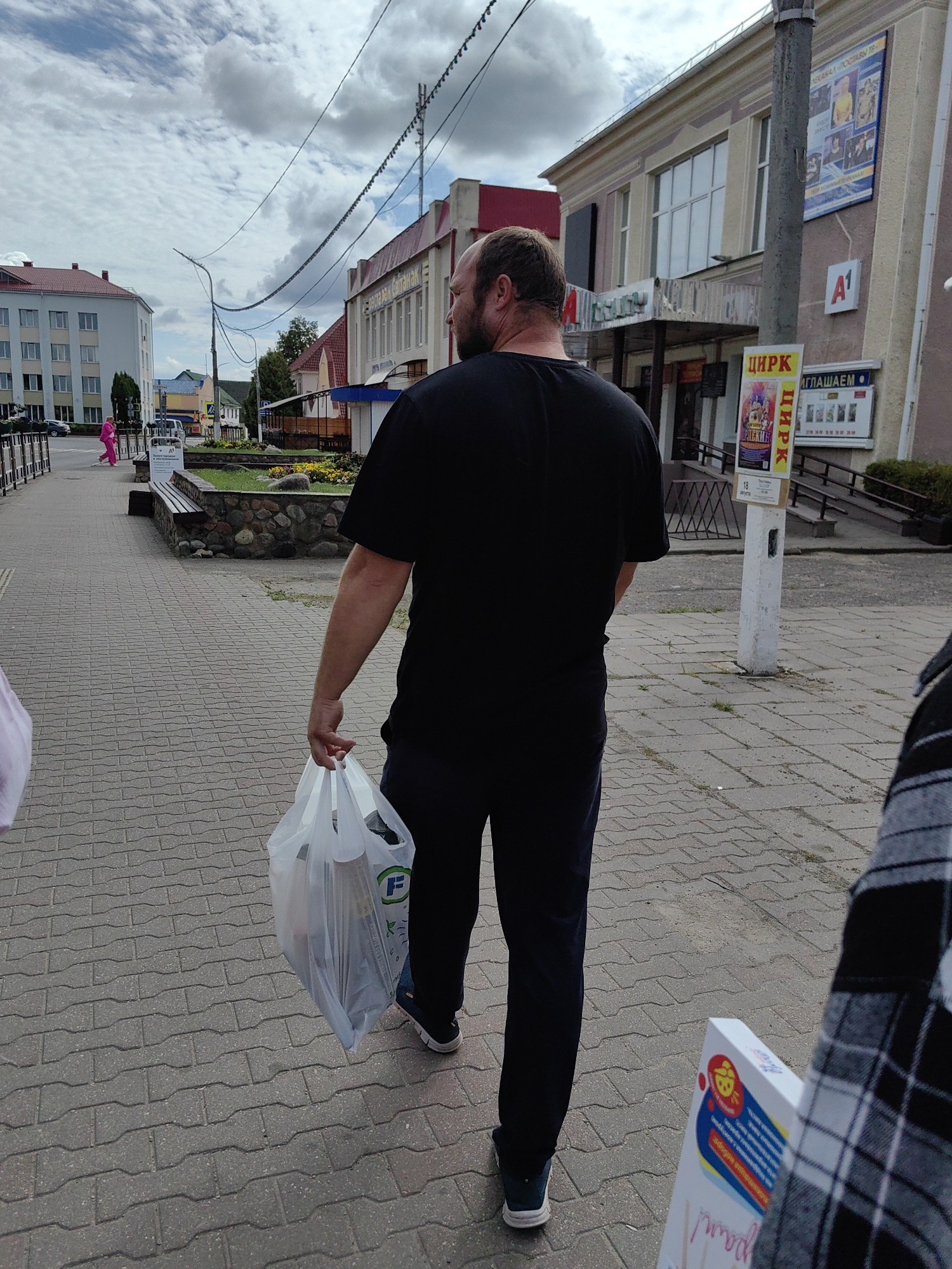 A bald man wearing a black t-shirt and dark pants walks away from the camera, carrying a white plastic grocery bag in his right hand. He is on a paved sidewalk in front of buildings. The building to his right has signs that include "ЦИРК" (circus) and "А1". A poster for a circus is visible on a pole near the building entrance. To the left of the man, another person's shoulder and arm, wearing a plaid shirt, are partially visible. In the background, there is a light-colored multi-story building and trees. The sky is cloudy. The location is the city of Pastavy, Belarus, on a street with a paved sidewalk and buildings lining one side.