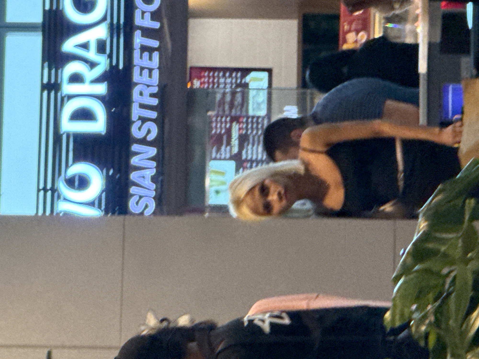 In Bắc Cam Ranh, Vietnam, a woman with blonde hair, wearing a black tank top, stands in front of a restaurant with a neon sign that reads "ASIAN STREET FOOD." The restaurant's menu is visible behind her. To her left, a person wearing a black hoodie with a pink shirt underneath stands partially obscured by the frame. To her right, the back of a man in a blue shirt is visible. In the foreground, a large green plant fills the bottom of the image.