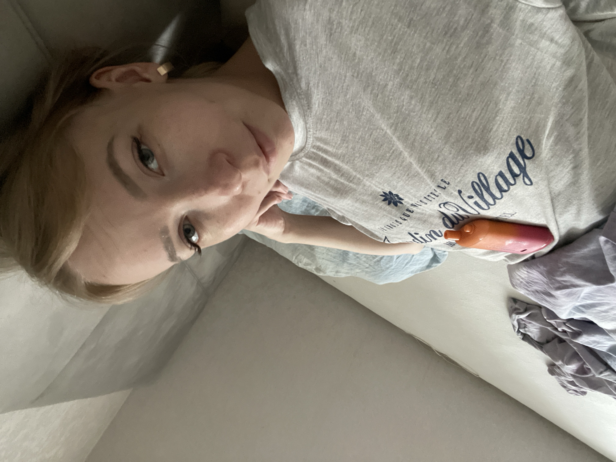 A fair-skinned person with blonde hair and light eyes is lying on a gray sofa. The person is wearing a light gray t-shirt with a blue design featuring the words "Notre Village" written on it. An orange-colored object is resting on the t-shirt near the design. Crumpled light-colored fabric is visible on the right side of the sofa. The setting appears to be indoors. There is no information available in the image to ascertain that this image was taken in Perm, Russia.