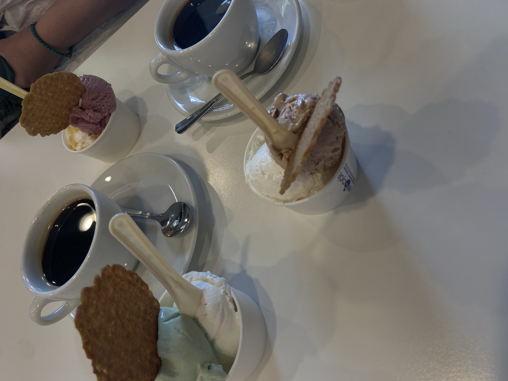 The image captures a table setting featuring three cups of gelato and two cups of coffee, likely taken at a café in Lecco, Italy. Each gelato cup contains two scoops of different flavors, topped with a wafer cookie and a plastic spoon. The coffee cups are white ceramic and sit on matching saucers, each with a spoon. The table is white. In the upper left corner of the image, part of a person's arm is visible. The overall composition suggests a casual cafe setting.
