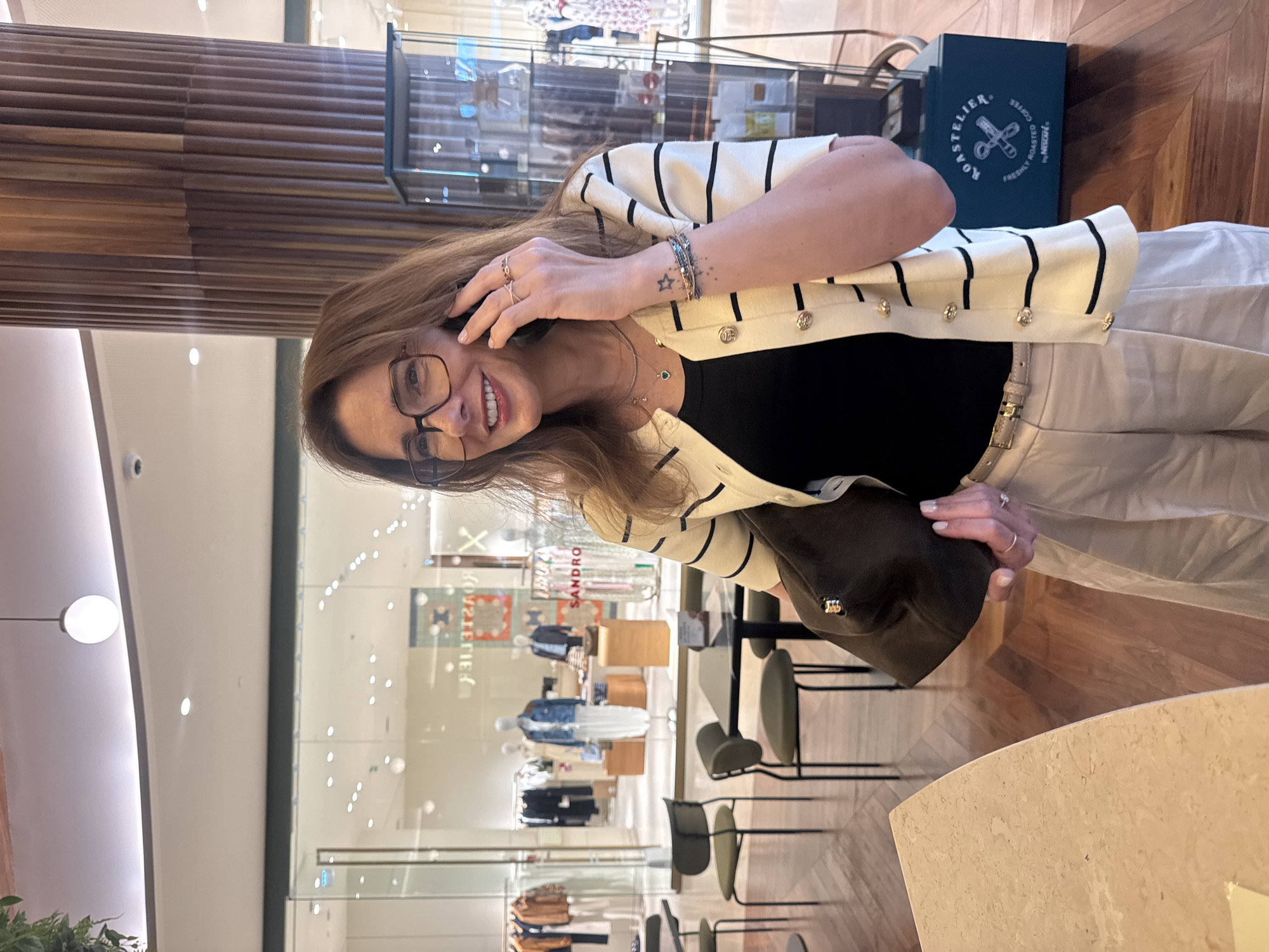 The image shows a woman indoors, possibly in a retail environment. She is wearing glasses, a black top, a white cardigan with black stripes, and light-colored pants. She holds a dark-colored hat or bag in one hand and appears to be on a phone call. In the background is a store with clothing displays visible through a glass window. The brand "SANDRO" is visible on the window. Furniture, including chairs, is also visible. A wooden column and a display case with a product box that reads "ROASTELIER COFFEE" can be seen as well. The setting appears to be a modern retail space. The location is Senhora da Hora, Portugal.