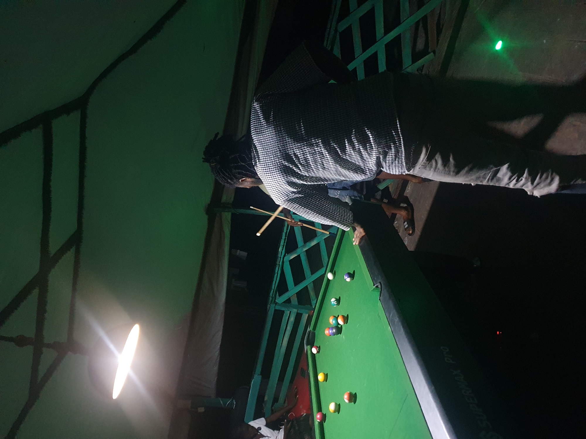 This is a full-shot, outdoor, low-light photograph of a billiard table at night, possibly in Lokoja, Nigeria. The scene appears to be in a somewhat rustic, open-air setting, suggested by the visible beams and tarpaulin overhead, and the green metal railings in the background. A single, bright overhead light illuminates the billiard table and the area around it.
In the foreground, a person with dreadlocks, wearing a checkered shirt and ripped jeans, is standing with their back to the camera, holding a billiard cue. They are positioned near the center of the table, appearing to be playing or about to play.
On the billiard table, several colored balls are scattered, including red, yellow, blue, orange, and a cluster of balls near the center. The table itself is green, with black edges. The text "SUPERMAX Pro" is visible on the side of the table.
In the background, another person wearing a white shirt is seated on a bench near the billiard table. Their posture suggests they are observing the game or waiting their turn. The environment is dark beyond the illuminated area, with the green railings of the seating area clearly visible.
Two small green lights are visible on the ground in the lower right corner, and a small red light is visible on the side of the table in the lower left. The overall impression is that of a casual, nighttime game of billiards in a semi-open, possibly entertainment-focused location.