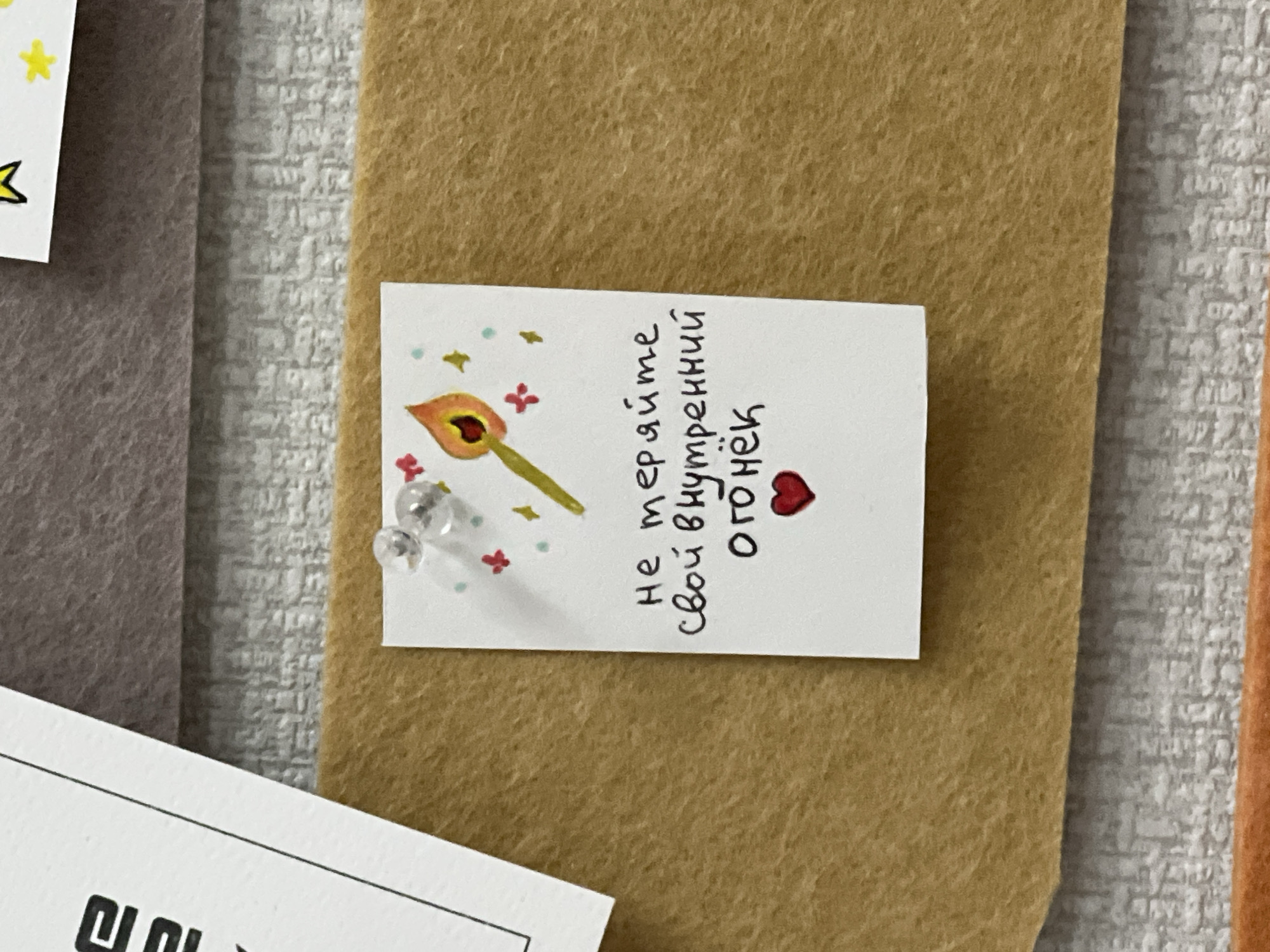 The image shows a close-up of a bulletin board with several pinned items. One prominent item is a small, rectangular white card pinned to a tan felt backing with two clear pushpins. The card has a drawing of a lit match, along with small star and cross-shaped decorations. Handwritten vertically on the card in black ink is the Russian phrase "не теряйте свой внутренний огонёк" and a small red heart. The text translates to "don't lose your inner fire." The surrounding background consists of a textured gray surface and other bulletin board items, including a gray felt backing and parts of white cards with writing or drawings.
The image does not provide enough details to determine the location in the city of Chaikovsky, Russia, or to identify any specific people, actions, or interactions.