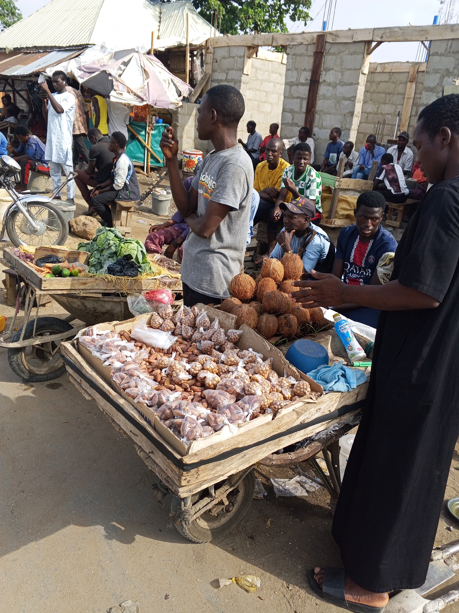 This image captures a vibrant outdoor market scene in Zango-ndaji, Nigeria, under bright daylight. The foreground features two men and two wooden pushcarts serving as market stalls.
On the right, a man in a black robe stands, his hands extended over a stall filled with various nuts and dried fruits, many packaged in small clear plastic bags. A prominent pile of coconuts rests on this same cart, alongside a blue plastic container and a bottle with a partially visible label that appears to read "NATURA" or "NATURAL" with a green and white logo. Facing him is another man in a grey t-shirt, looking towards the right. His shirt bears the text "GRAPHIC".
To the left, another pushcart displays fresh produce, including a large mound of green leafy vegetables (likely cabbage or lettuce), carrots, limes, and eggplants. The front wheel and handlebars of a motorcycle are visible next to this cart.
In the mid-ground and background, numerous other men are gathered. Some are sitting on benches or the ground, observing the market activity or socializing. One man in a white tunic on the far left is looking at his phone. Visible clothing includes a blue and red football jersey with "QATAR AIRWAYS" printed on it, and a green and white striped shirt with an "adidas" logo. The market is set against makeshift shelters with corrugated iron roofs and tarpaulin awnings, and a partially constructed building with concrete block walls.
Strong shadows on the ground indicate a sunny day, likely mid-morning or afternoon. The overall atmosphere suggests a casual, bustling community market.