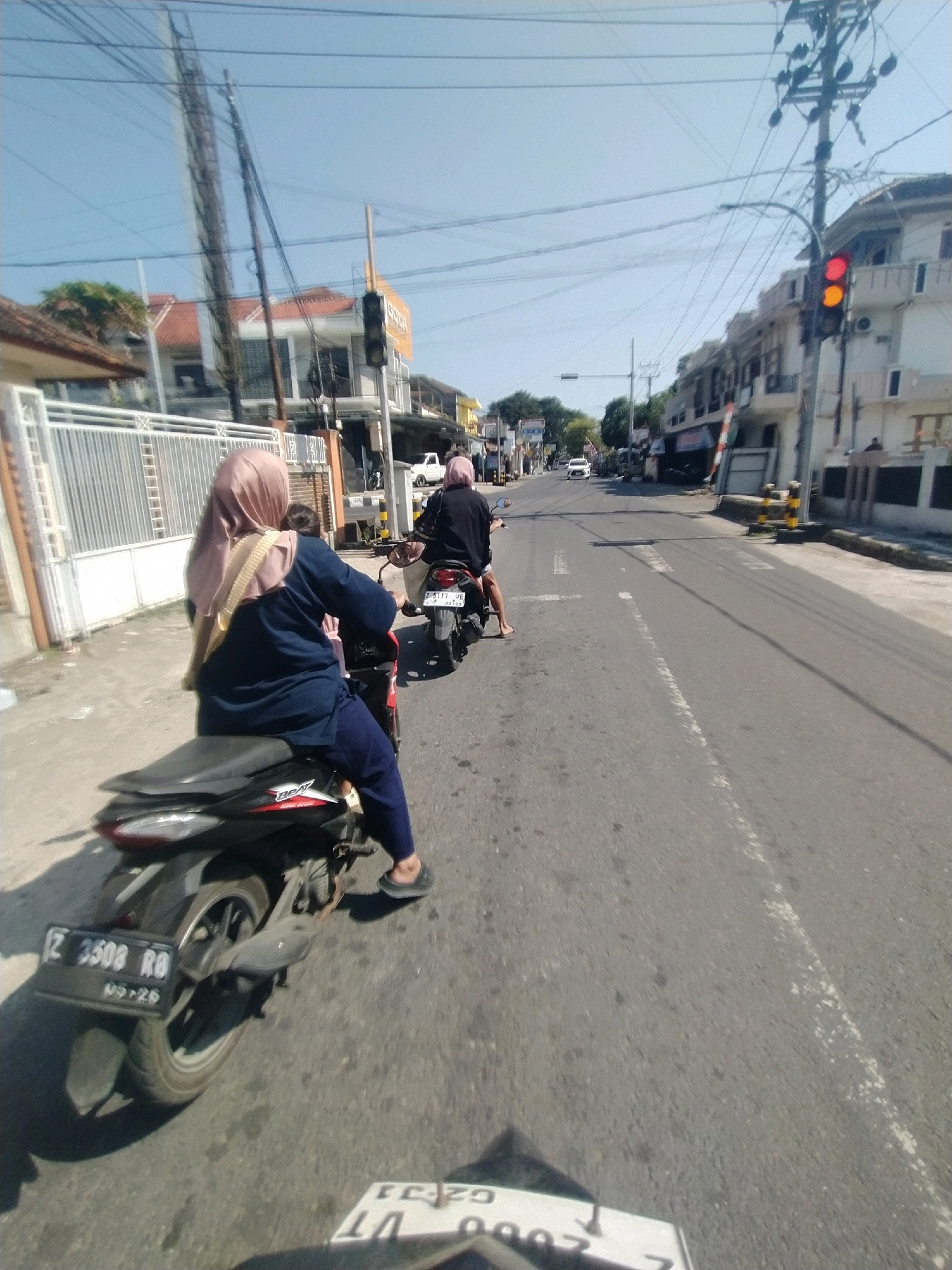 On a sunny day, two individuals are riding motorcycles in Desa Karangbenda, Indonesia. The lead motorcycle, a black and silver model, is carrying a person wearing a navy blue long-sleeved shirt, dark pants, and a light pink hijab. This rider is also wearing a beige strap over their left shoulder. The second motorcycle, also black, is following behind. The street is paved and marked with white lines. Buildings with tiled roofs line the right side of the street, with power lines overhead. A traffic light is visible on the right, showing red and yellow lights. A white car is visible in the distance on the left side of the road.