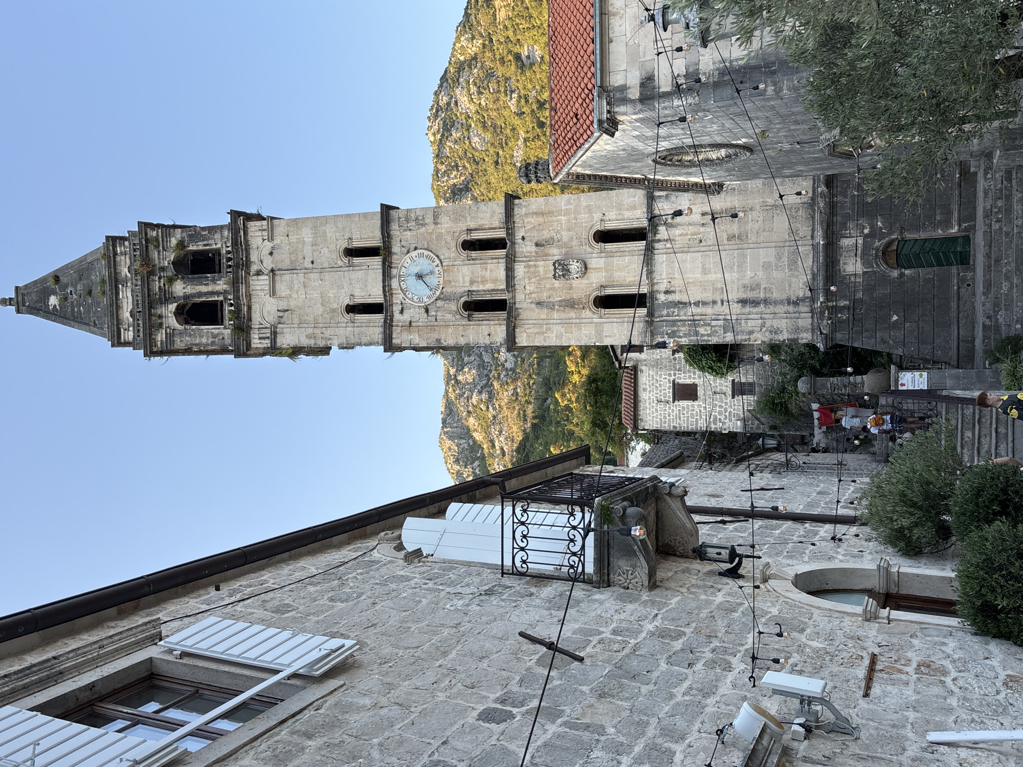 The image shows a view of the bell tower of St. Nicholas Church in Perast, Montenegro. The stone bell tower, which dominates the right side of the frame, is tall and slender with arched openings and a conical spire. A large clock face is visible on one side of the tower. To the left of the tower is a stone building with a balcony and white shutters. String lights with exposed bulbs hang across the narrow street. In the background, a rocky, tree-covered hillside rises beneath a clear blue sky. Several people are seen walking up a stone staircase leading away from the camera, towards the church. The scene appears to be set in a historic European town with traditional architecture.