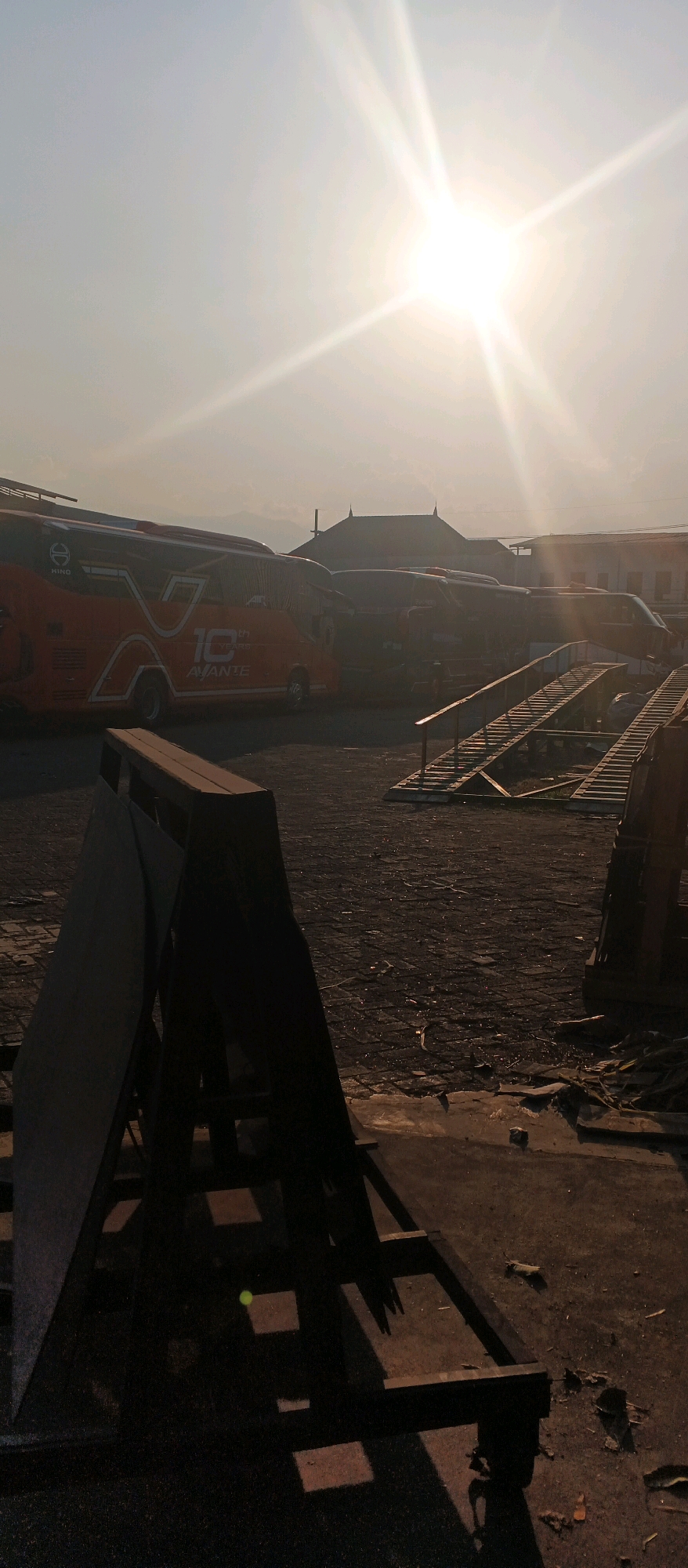 The image captures a scene at an outdoor parking lot or bus depot, likely in Banjararum, Indonesia, during a bright, sunny day with the sun low in the sky, casting long shadows. In the foreground, a dark, angular metallic structure, possibly a ramp or a stand, dominates the left side of the frame. Beyond this, the ground appears to be a mix of asphalt and gravel, with debris scattered across it.
Several buses are parked in the mid-ground. The most prominent is a large, orange bus with white and black accents, displaying "HINO" and "10th AVANTE" text. Other buses, partially obscured by shadows and distance, are also visible. In the background, there are low-rise buildings with dark roofs, one featuring a distinctive pitched roofline. The sky is bright and hazy, with the sun appearing as a powerful, starburst-like light source, creating lens flare and washing out parts of the sky. The overall atmosphere is that of a working depot, possibly at the beginning or end of the day given the low sun angle and light conditions. No people are visible in the frame.