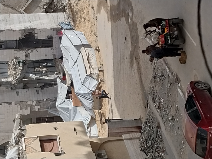 This image depicts a street scene in Khan Yunis, Palestinian Territory, characterized by significant urban destruction and makeshift living arrangements. In the background, a large, multi-story building, likely a former residential or commercial structure, stands heavily damaged with substantial portions of its facade missing, exposing internal debris and structural elements.
In the midground, an expanse of sandy, uneven terrain serves as a temporary settlement, occupied by numerous white canvas tents, several of which display "UNRWA" branding. Individuals, including children, are present within this camp area; one adult male is observed carrying a small child while moving across the sandy ground. Another person is visible near the doorway of a building on the far left.
The foreground features a paved road, partially covered by debris and dirt. A red Hyundai i20 sedan is parked on the left side of this road. On the right, three adult males are gathered around a four-wheeled pushcart laden with various fruits and vegetables. One man in dark clothing is bent over a cardboard box on the ground adjacent to the cart, while the other two stand upright nearby. Piles of rubble and excavated earth are visible along the road's edge. A portion of a beige building with a decorative window frame and an external air conditioning unit is visible on the far left.