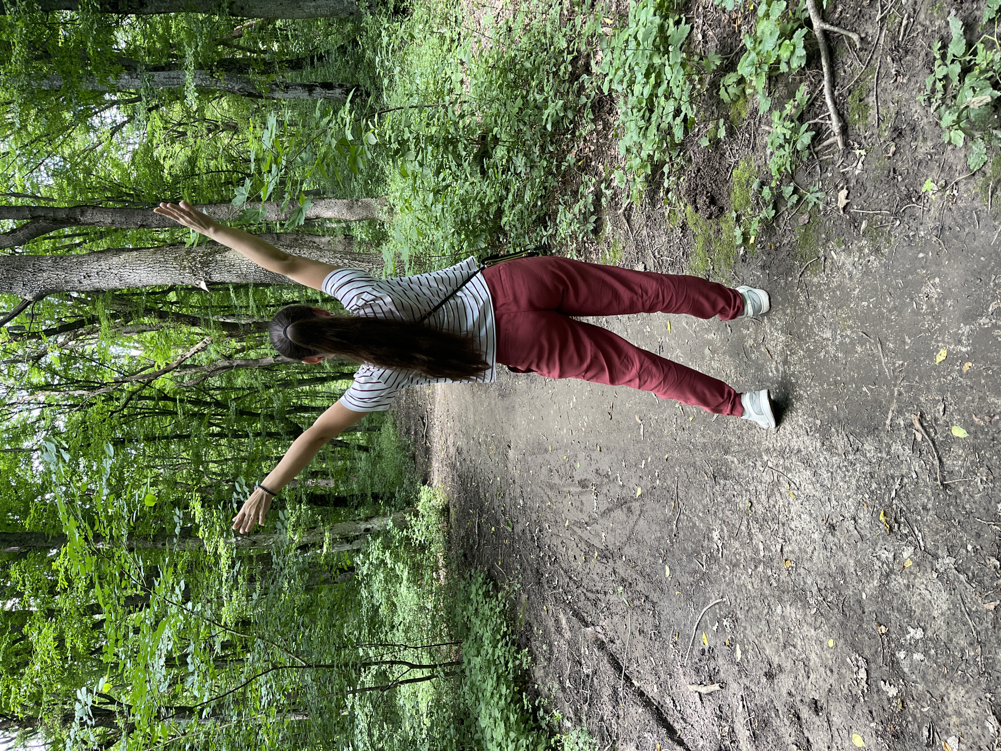 A person is performing a side plank exercise on a dirt path in a forest. The individual, facing away from the camera, has long dark hair and is wearing a striped t-shirt and maroon pants. Their arms are extended outwards, parallel to the ground, supporting their body. The setting is a lush forest with dense green foliage and trees lining a well-worn path. Sunlight filters through the leaves, indicating it is daytime, and the weather appears clear. The ground is a mix of dirt, fallen leaves, and small branches. There are no visible text elements in the image. The overall mood suggests a peaceful, active moment in nature.