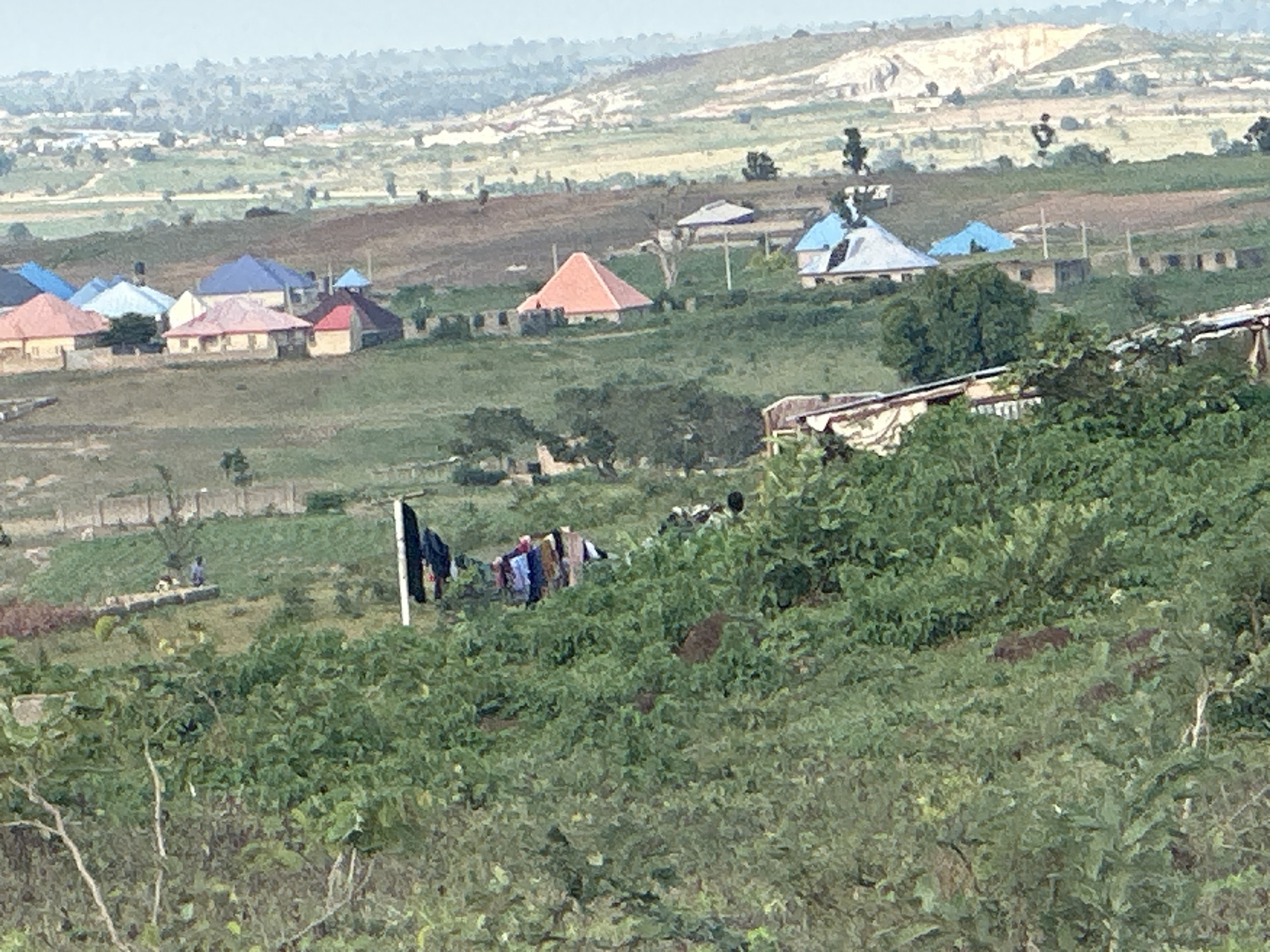 The image depicts a hilly landscape with scattered residential buildings and vegetation in the city of Gidan Waziri, Nigeria. In the foreground, a dense area of green foliage partially obscures the view of the scene. A clothesline with several items of laundry is visible on the left side, with a dark flag or banner also present. A person in blue clothing stands near a concrete structure in the middle ground.
Further into the scene, a cluster of houses with various colored roofs, including blue, pink, and red, are situated on the hills. One house features a prominent orange conical roof. In the background, the landscape transitions to lighter green and brown hills, with evidence of quarrying or excavation at the top. The overall atmosphere suggests a rural or semi-rural setting within a developing urban area.