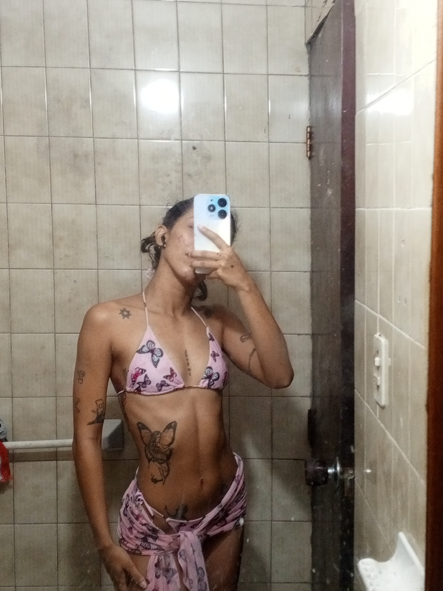 A person with tattoos is taking a selfie in a bathroom with tiled walls. The person is wearing a pink bikini top with a butterfly pattern and a matching sarong tied around their waist. They are holding a smartphone with a blue case in their right hand, obscuring their face. The bathroom has a dark wooden door and a mirror. The condition of the tiles and the door suggests an older, possibly humid environment. There is no readily apparent information in the image to verify the location in the city of Porlamar, Venezuela.
