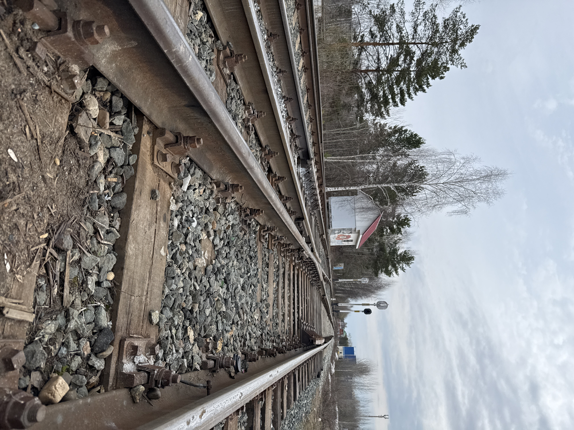 The image shows a set of railroad tracks extending into the distance. The tracks are bordered by gravel and secured to wooden ties with metal fasteners. In the background, there are trees, and a small white building with a red roof is visible. Further along the tracks, railroad signals can be seen. The sky is cloudy. The location is Unknown, Unknown.