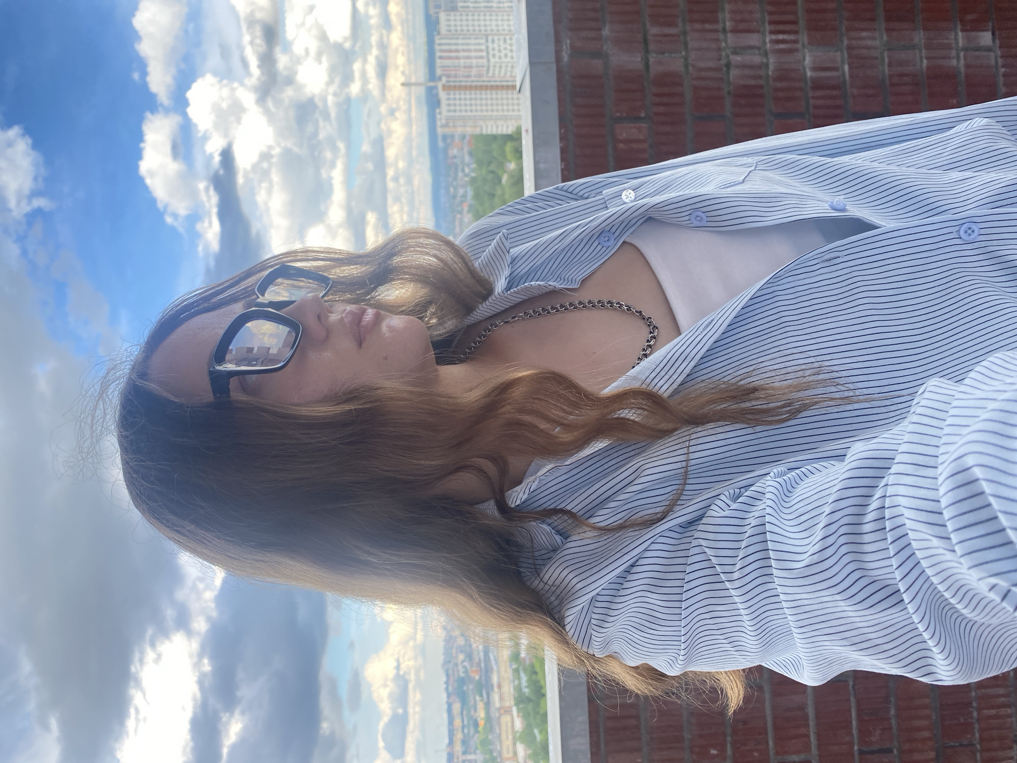 A young woman wearing glasses and a striped blue shirt poses on a rooftop in Izhevsk, Russia. She is also wearing a silver necklace and a white undershirt. Behind her, city buildings are visible in the background, along with a cloudy sky. A brick structure is also present at the right of the image. The photo appears to have been taken from a low angle, looking upwards.
