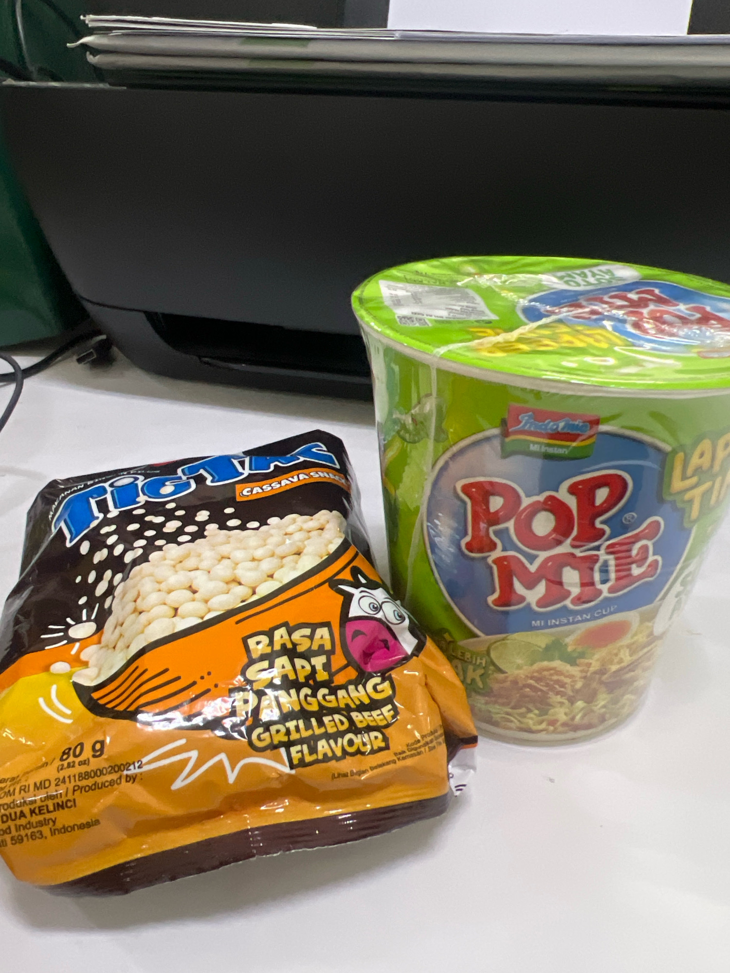 Two snack items, a bag of Tigis Cassava Snacks and a cup of Pop Me Mi Instan, are placed on a white surface, likely a desk or table, in front of a black office printer. The Tigis bag is brown and orange, featuring an illustration of white cassava snacks and text including "Rasa Sapi Panggang Grilled Beef Flavour." The Pop Me cup is green and blue, with "Pop Me" prominently displayed. The background shows the upper part of the black printer and stacks of paper. The scene appears to be in an indoor setting, possibly an office, and there is no indication of its specific location within Yogyakarta, Indonesia.