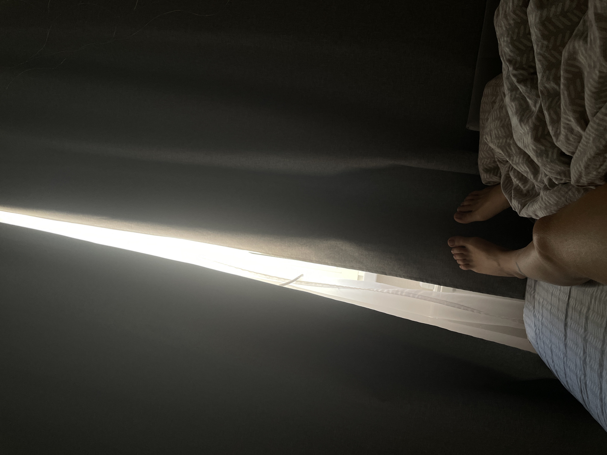 This image depicts a person's bare feet and lower legs peeking out from under a patterned blanket on a bed. The person is positioned on the right side of the frame, with their feet facing upwards and to the left. A bright light streams in from the left side of the image, partially obscured by dark curtains that are drawn open. The light illuminates a sheer white curtain behind the dark curtains, and the faint outline of a window can be seen through it. The environment suggests an indoor setting, likely a bedroom, with the bed occupying the foreground and the bed sheets having a grey and white patterned design. The overall lighting is dim, with the bright light from the window creating a strong contrast. The image does not contain any discernible features or elements that would allow for the determination of a specific location within Rostov-on-Don, Russia.