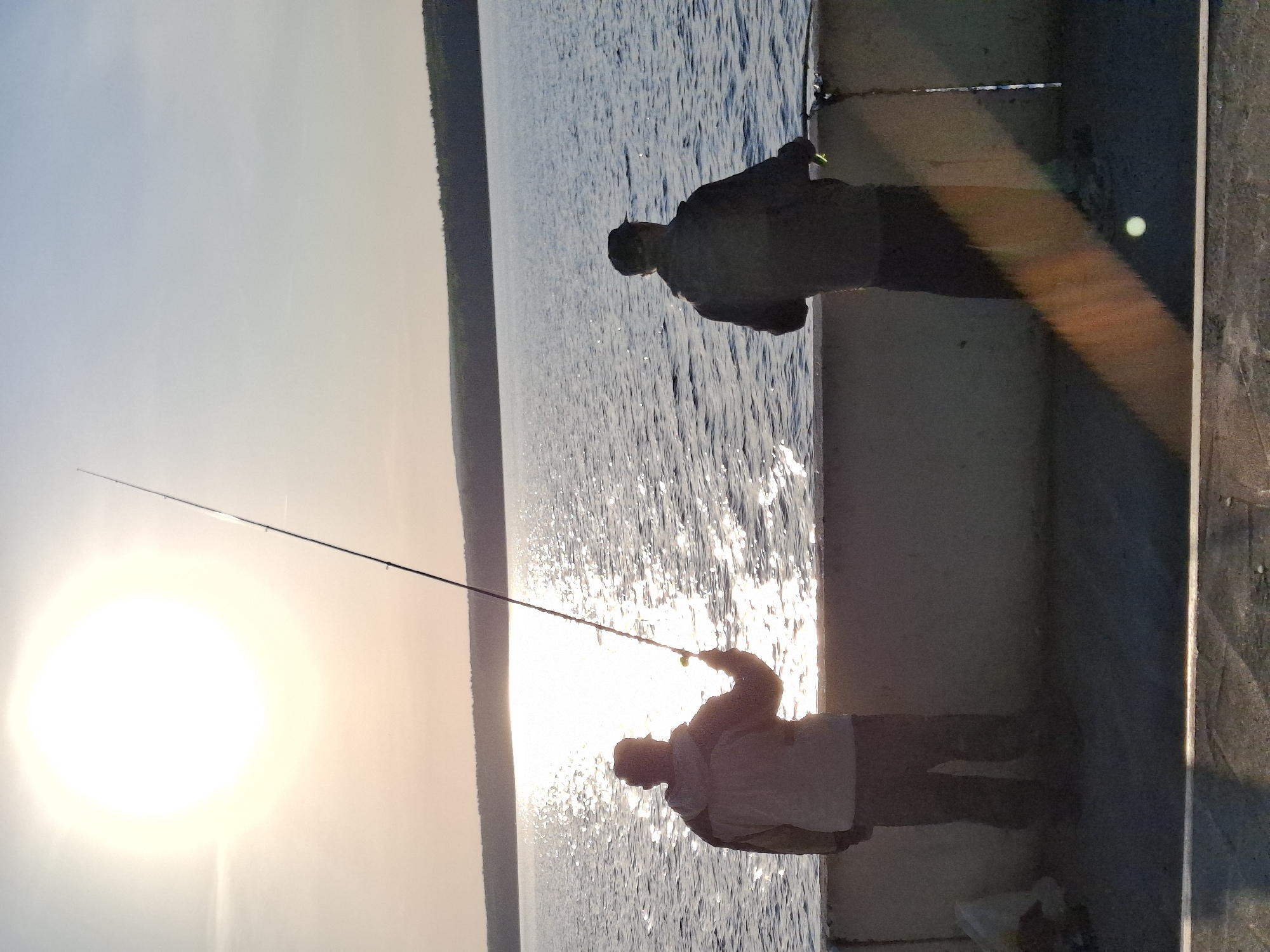 Two people are fishing from a concrete pier. The bright sun is low in the sky, possibly at sunrise or sunset, creating silhouettes of the fishermen. A body of water extends to the horizon. Tree-covered land is visible in the distance. The location is Chaikovsky, Russia.