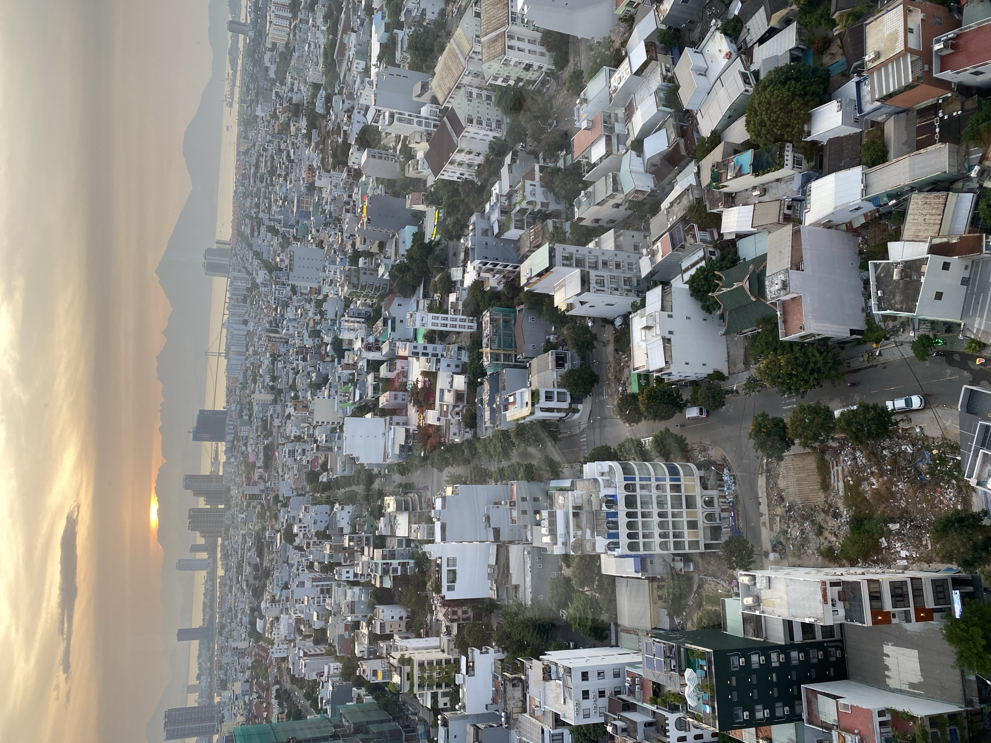 The image shows a panoramic view of the city of Da Nang, Vietnam, from a high vantage point, likely a building. The time of day appears to be sunset or sunrise, as evidenced by the warm, hazy light in the sky and the low sun peeking through the clouds. In the distance, mountains form a backdrop to the cityscape and the sea. A prominent bridge spans the water.
The foreground and midground are densely packed with buildings of varying heights and architectural styles, characteristic of a rapidly developing urban area. Many buildings are residential, with a mix of apartment complexes and smaller houses. Some buildings are under construction, indicated by scaffolding and exposed frameworks. The streets are narrow and winding, with a few cars and motorcycles visible.
There are areas of green space, primarily trees and some smaller parks, interspersed among the buildings. However, one area in the mid-foreground is notably filled with a large amount of debris and refuse, suggesting a neglected or unmanaged lot. This contrasts with the generally urban environment.
The overall impression is of a bustling, growing city with a mix of modern development and some signs of urban challenges. The natural beauty of the surrounding landscape is visible in the distance, providing a scenic element to the urban sprawl.