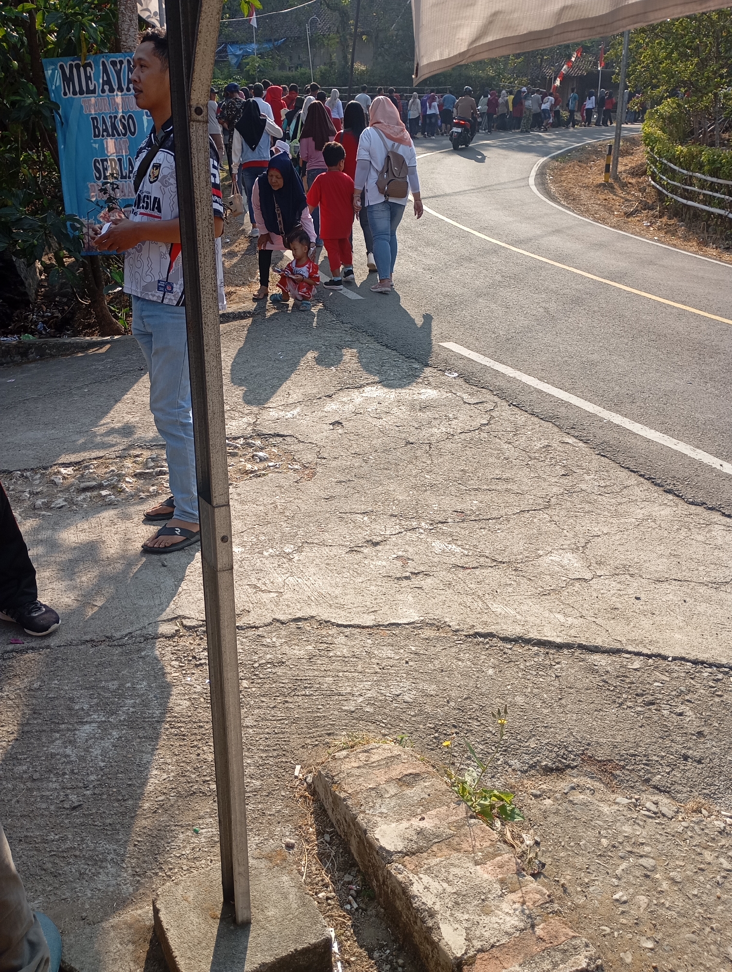 The image depicts a street scene in Ketepung, Indonesia. In the foreground, a man wearing a white and black patterned shirt and blue jeans stands near a blue banner advertising "MIE AYAM" (Chicken Noodles) and "BAKSO" (Meatballs). To his right, a group of people, including women in hijabs and children, are walking along the paved road. Some are dressed in red and pink clothing. A motorcycle is visible further up the road, and more people are gathered on the side. The environment is sunny, with trees and foliage on the left and right sides of the road. The road is asphalt with yellow and white markings. The ground in the foreground is cracked concrete with some vegetation growing through it.