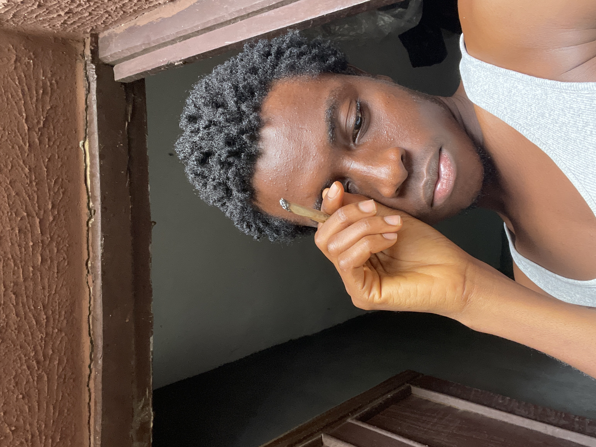 This is a close-up, vertically oriented photograph of a young Black man. He is wearing a grey tank top and has short, dark, curly hair. His face is turned to the side, with his eyes looking downwards. He is holding a rolled cigarette between the thumb and forefinger of his right hand, bringing it close to his face. His skin appears smooth and slightly shiny.
The background is dark and out of focus, suggesting an indoor setting or late evening. To the left of the frame, a textured, reddish-brown surface is visible, possibly a wall or doorframe. This is juxtaposed with darker brown wooden elements at the top and bottom of the image, which could be part of a window or doorway.
The lighting in the image is soft, casting gentle shadows and highlighting the contours of the man's face and hand. His expression appears thoughtful or perhaps melancholic. There is no visible text in the image. The overall mood is intimate and introspective. The location context is Nkpor, Nigeria, though no specific landmarks are identifiable within the frame.