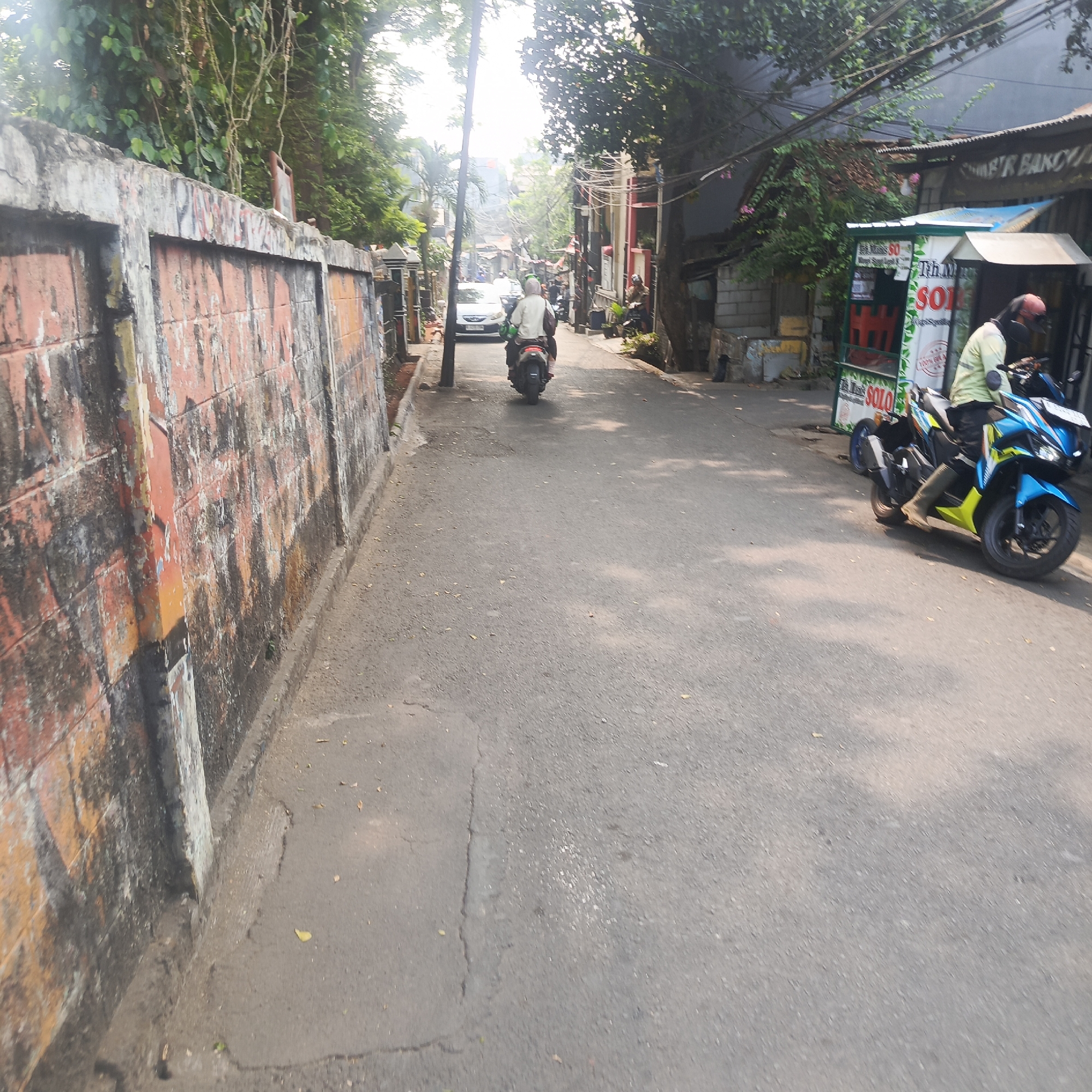 In this scene, a narrow paved street in South Jakarta, Indonesia, is depicted. The street is lined with buildings and vegetation. On the left, a weathered, red brick wall with graffiti runs alongside the road. On the right, a small food stall with a blue and green sign reading "THM SOLO" is visible. A person wearing a light green shirt and a red helmet is riding a blue and yellow motorcycle past the stall, moving away from the viewer. Further down the street, another motorcycle with two riders, one wearing a white shirt and a green helmet, is also moving away. A white car is also visible in the distance. The sky is bright and sunny, with some trees and power lines visible. The overall impression is of a busy, urban environment with everyday activities taking place on the street.