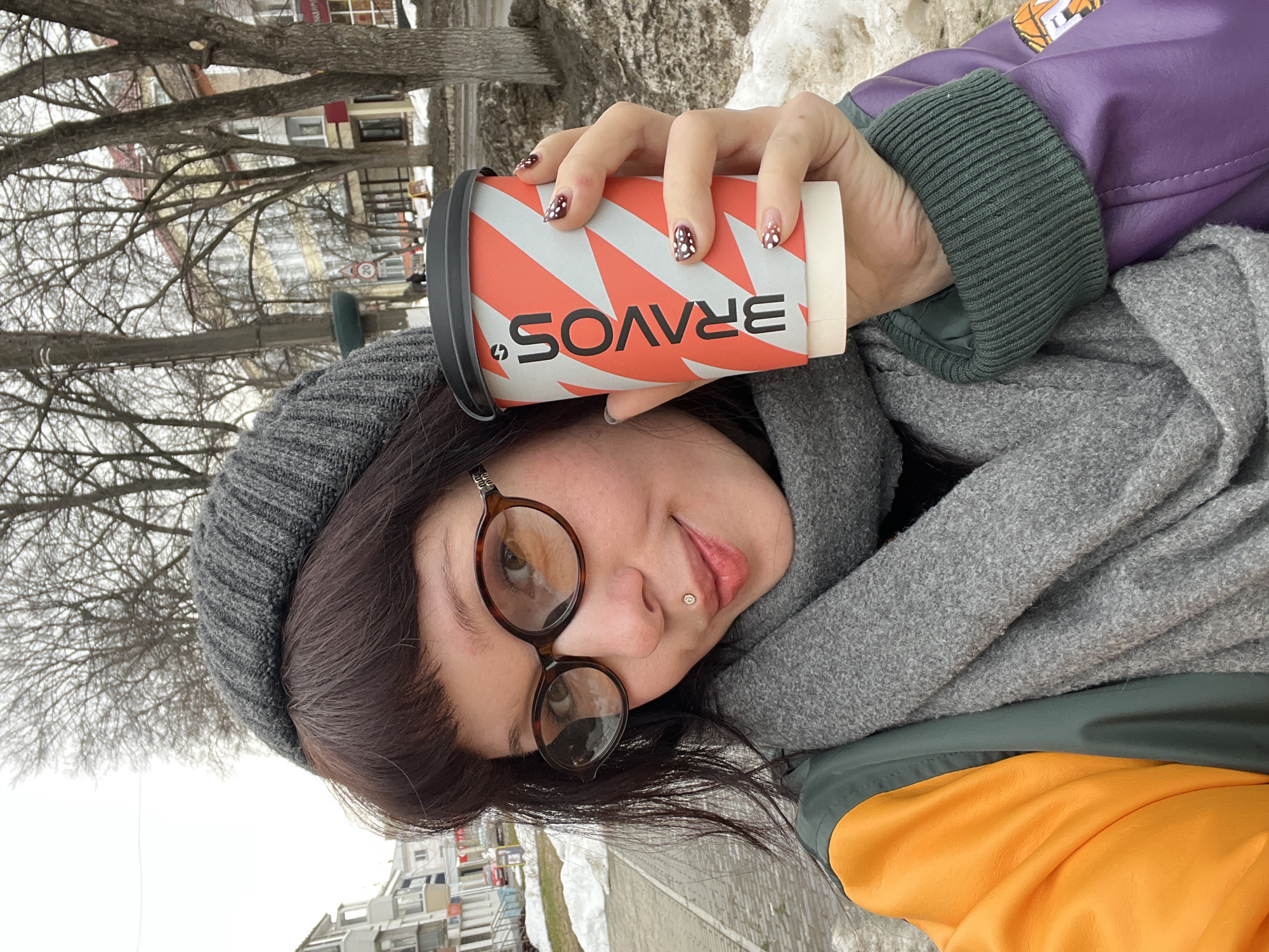 A female subject is depicted in a close-up, selfie-style portrait, positioned outdoors on a city street in Chaikovsky, Russia, during what appears to be late winter or early spring. She has dark brown hair with bangs, wears brown oval-framed glasses, and a small gem-studded labret piercing on her lower lip. Her attire includes a grey ribbed knit beanie, a grey knitted scarf, and a multi-colored jacket featuring distinct orange, dark green, and purple sections with a green ribbed cuff on the right sleeve. She holds a white and orange paper coffee cup with a black lid in her right hand, displaying "BRAVOS" printed vertically in black. Her fingernails are manicured with dark maroon and white polka dot patterns. The background reveals a paved sidewalk with patches of melting snow on the adjacent ground. Bare deciduous trees line the street. Across the street, multi-story buildings are visible, including a light-colored residential structure on the left and a building with a yellow facade, red trim, and ground-level shops on the right. A street sign indicating "20" is faintly discernible. The sky is overcast.