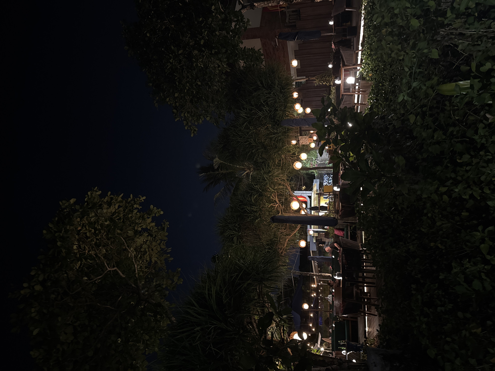 This image depicts an outdoor dining area at night, likely a restaurant or bar, in Kuta, Indonesia. The scene is illuminated by numerous round, orange-toned string lights hanging from trees and poles, casting a warm glow on the surroundings. Several tables with chairs are visible, some occupied by patrons. Green foliage and trees frame the foreground and midground, creating a sense of natural ambiance. In the background, structures with visible signage and lighting suggest the presence of buildings and other businesses. The sky is dark, indicating it is nighttime.