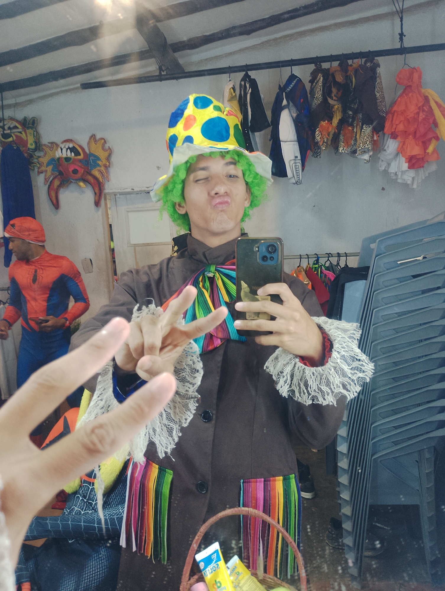 A person dressed as a clown takes a selfie in a mirror. The clown has green hair, a yellow and blue polka-dot hat, a brown jacket with rainbow fringe, and ruffled cuffs. The clown is making a kissy face and holding up two fingers in a peace sign. In the background, a person in a Spider-Man costume is visible. Other costumes hang on a rack behind the clown. A pile of stacked chairs is on the right side of the image. Additional details include masks hung on the wall and what appears to be a storage area, possibly in Porlamar, Venezuela, given the prompt's context.