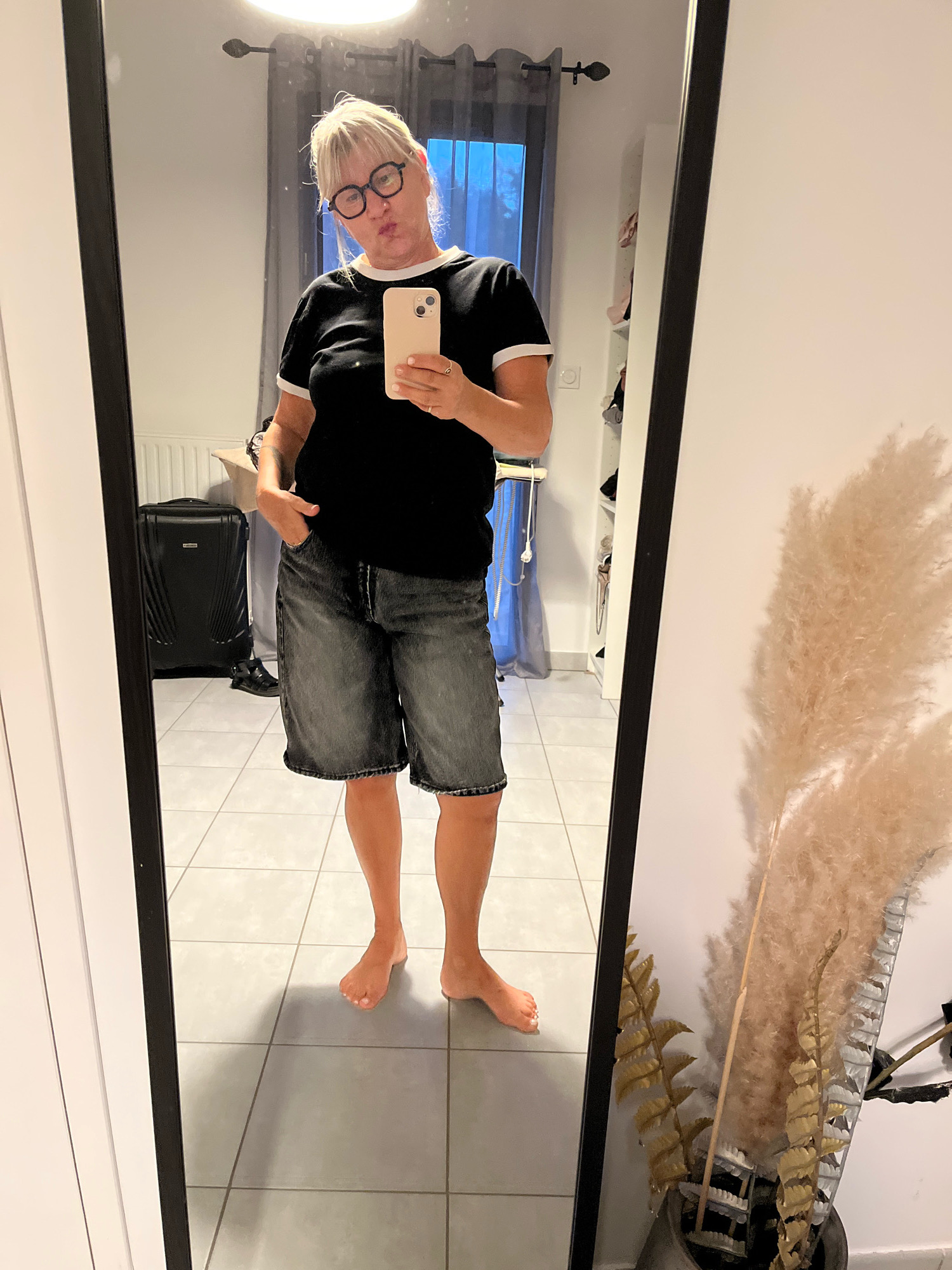 A person is standing in front of a mirror, taking a selfie. They are wearing a black t-shirt with white trim and denim shorts. The person has blonde hair and is wearing glasses. In the background, there is a suitcase, a window with curtains, and a vase with dried plants. The floor is tiled. The image was taken in Chambœuf, France.