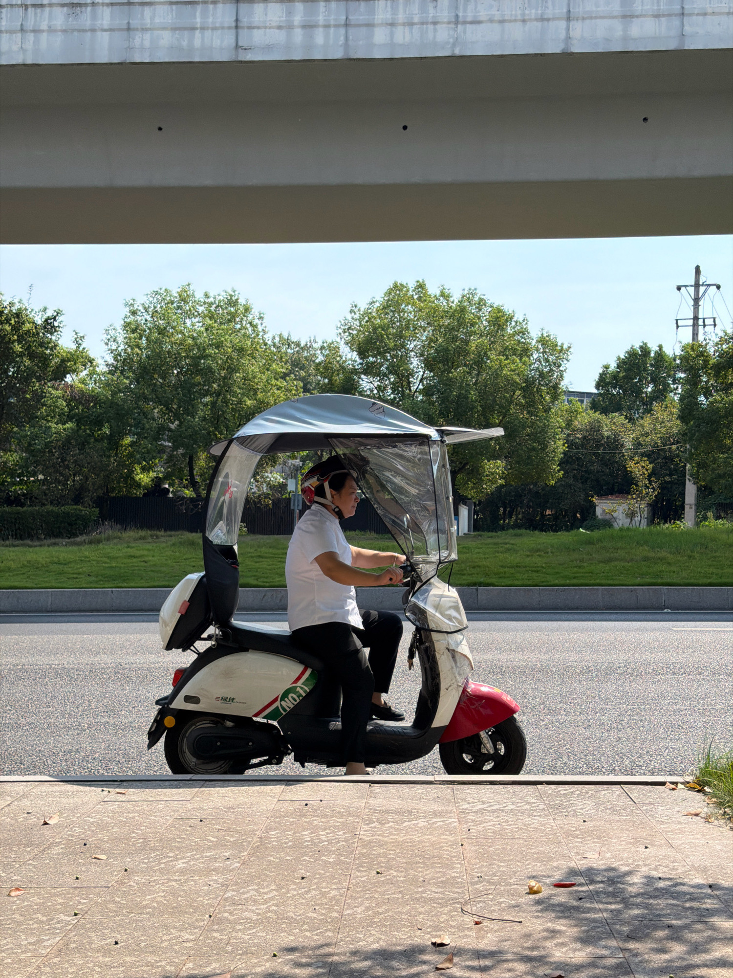 A person wearing a white polo shirt and black pants is riding a red and white electric scooter. The scooter has a transparent canopy and a black storage box on the back. The person is wearing a helmet and is holding the handlebars. The scooter is on a paved road next to a sidewalk. In the background, there are trees and a power pole. The scene is under an elevated concrete structure. The location is the city of Jiangjin, China.