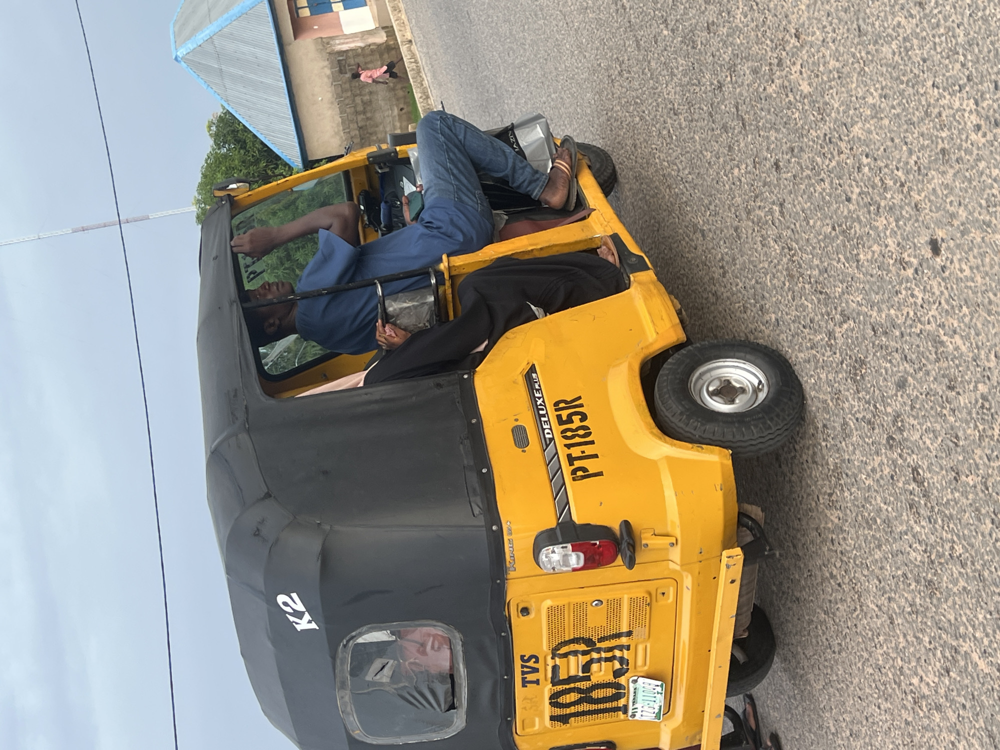 The image depicts a yellow and black TVS "King K2 Deluxe Plus PT-185R" tricycle, a type of motorized vehicle commonly used for transportation in Nigeria, parked on an asphalt road. The license plate reads "4851" and "TZG-11106A 11 IBADAN NIGERIA." Two individuals are visible within the tricycle; the driver is a black man wearing a blue shirt, while the passenger is also black and is dressed in black trousers. The location is possibly Howayi, Nigeria, based on the license plate information. A building with a blue roof is partially visible in the background, along with a pedestrian on the sidewalk.