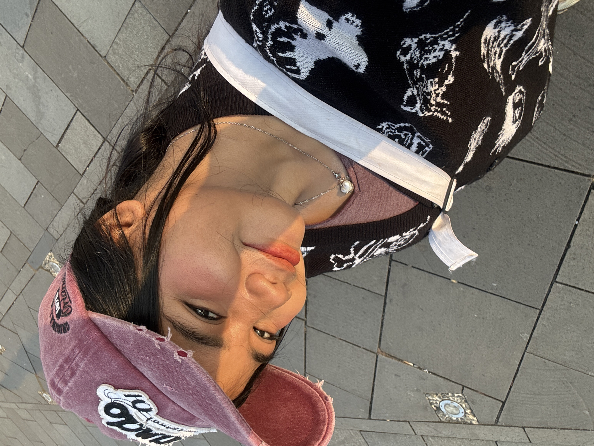 The image is a close-up, tilted selfie of a young woman wearing a pink distressed baseball cap with patches and a black sweater with a white pattern. She is looking towards the camera with a slight smile. Golden hour sunlight illuminates her face from the side, highlighting her cheekbones and lips. A silver necklace with a heart-shaped pendant is visible. She appears to be outdoors, with a grey tiled ground in the background. The scene suggests a casual, relaxed setting, possibly in a public area of West Jakarta, Indonesia, such as a park or pedestrian walkway.
