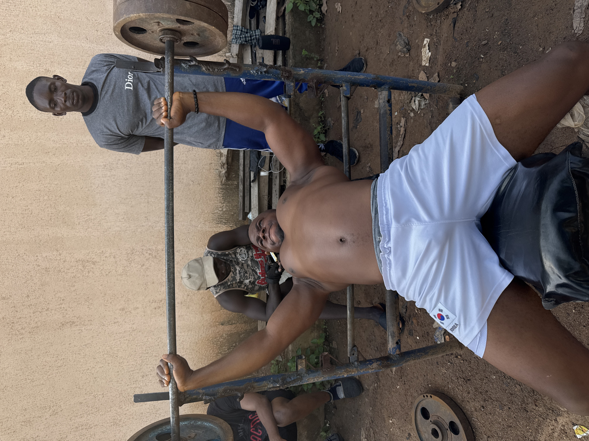 In an outdoor setting with a rough, dirt ground, three men are engaged in a workout session. The scene appears to be a makeshift gym, evident from the rustic metal weightlifting equipment. One man, wearing a grey t-shirt with "Dior" printed on it, stands to the left, holding onto a weightlifting bar. He has a serious expression on his face. In the center, another man, shirtless and wearing white shorts with a Korean flag emblem and the word "KOREA" visible, is lying on a bench, performing a bench press. His muscles are engaged as he lifts the weight. A third man, wearing a camouflage tank top and a light-colored cap, is positioned behind the man on the bench, observing the workout. The lighting suggests it is daytime, and the open-air environment indicates warm weather. The overall atmosphere is one of physical exertion and training.