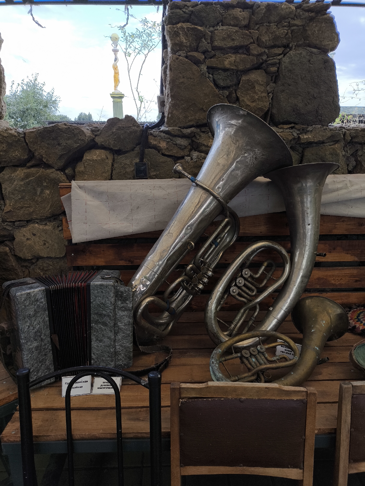 In Borgustanskaya, Russia, an image captures a collection of brass musical instruments and an accordion displayed on wooden planks against a stone wall. The instruments include a large tuba, a baritone horn, and a smaller brass horn, all showing signs of age and use. An accordion with a marbled grey finish rests to the left. In the background, through a window in the stone wall, a statue of a woman holding a globe on a pedestal is visible, along with trees and a cloudy sky. Two white signs with Cyrillic text are placed on the wooden surface in front of the accordion, and part of a chair is visible in the foreground. The scene suggests a display or storage area for musical instruments, possibly in an outdoor or rustic setting.