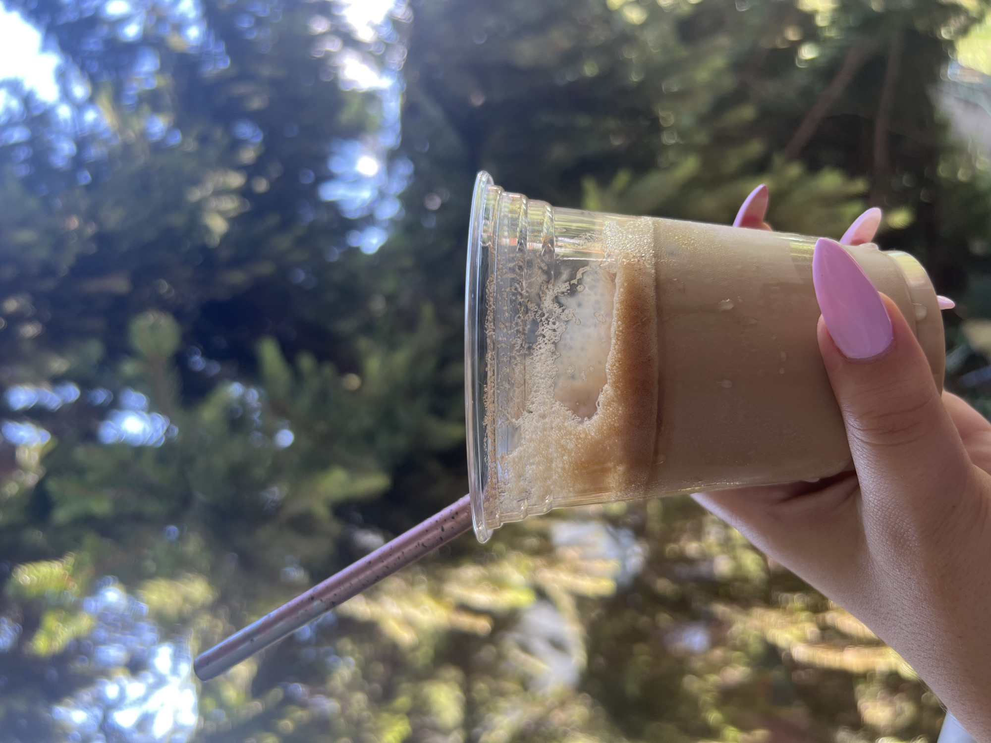A hand with pink painted nails holds a clear plastic cup filled with a frothy, light brown beverage. A metallic pink straw is inserted into the cup. The background is blurred, showing green foliage and sunlight filtering through. The image was taken in Sofia, Bulgaria.