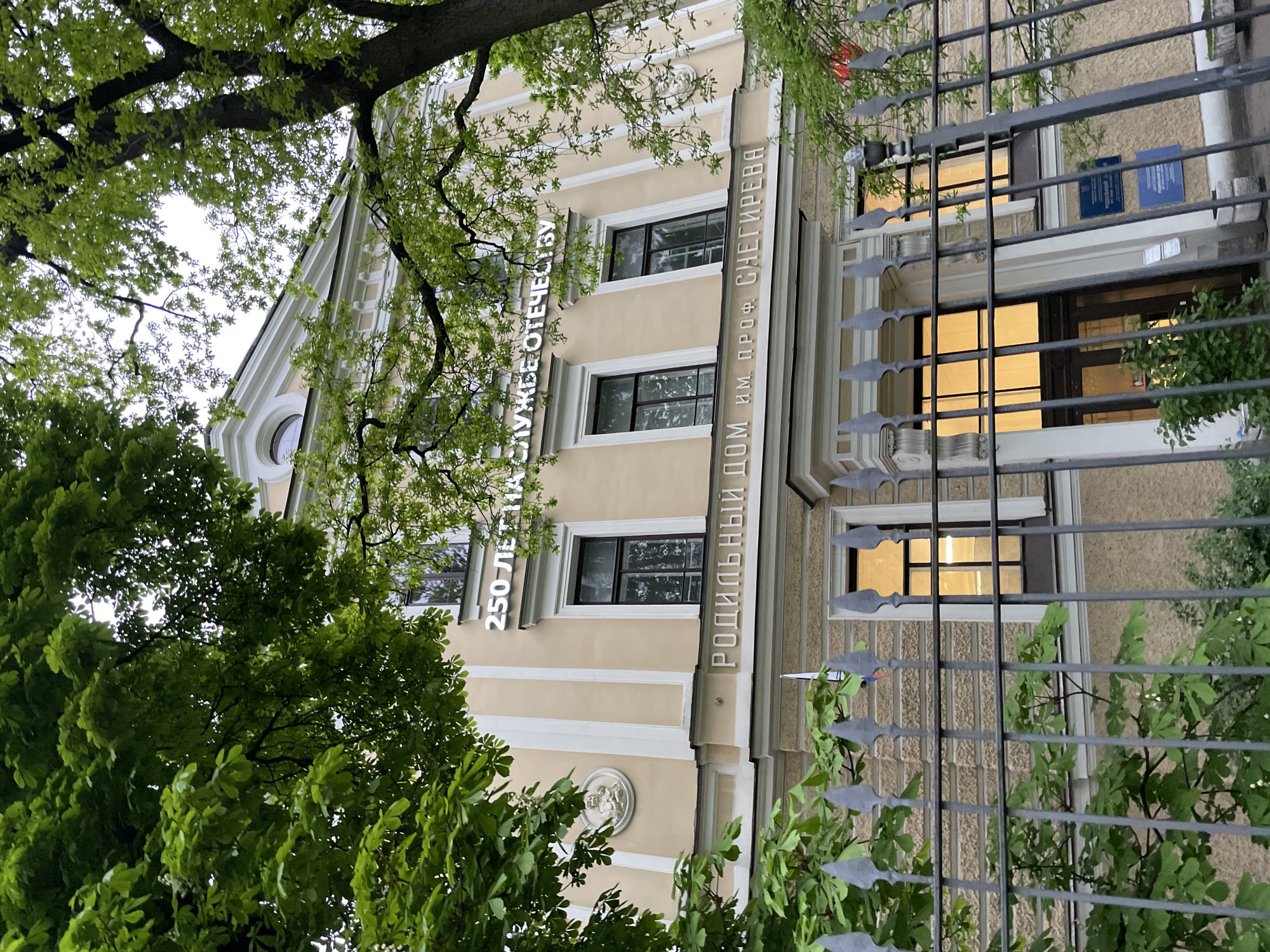 The image shows a building partially obscured by trees and a metal fence, likely in Saint Petersburg, Russia. The building is beige with white trim around the windows and along the vertical lines of the facade. The signage on the building is in Russian. One inscription reads "250 LET SLYZHBE OTECHESTVU" and the other says "RODIL'NIY DOM IM. PROF. SNEGIREVA". The second inscription suggests that the building is the Snegirev Maternity Hospital. The windows are dark, but there is light coming from inside. The metal fence is grey, and the background is somewhat blurred by the trees.