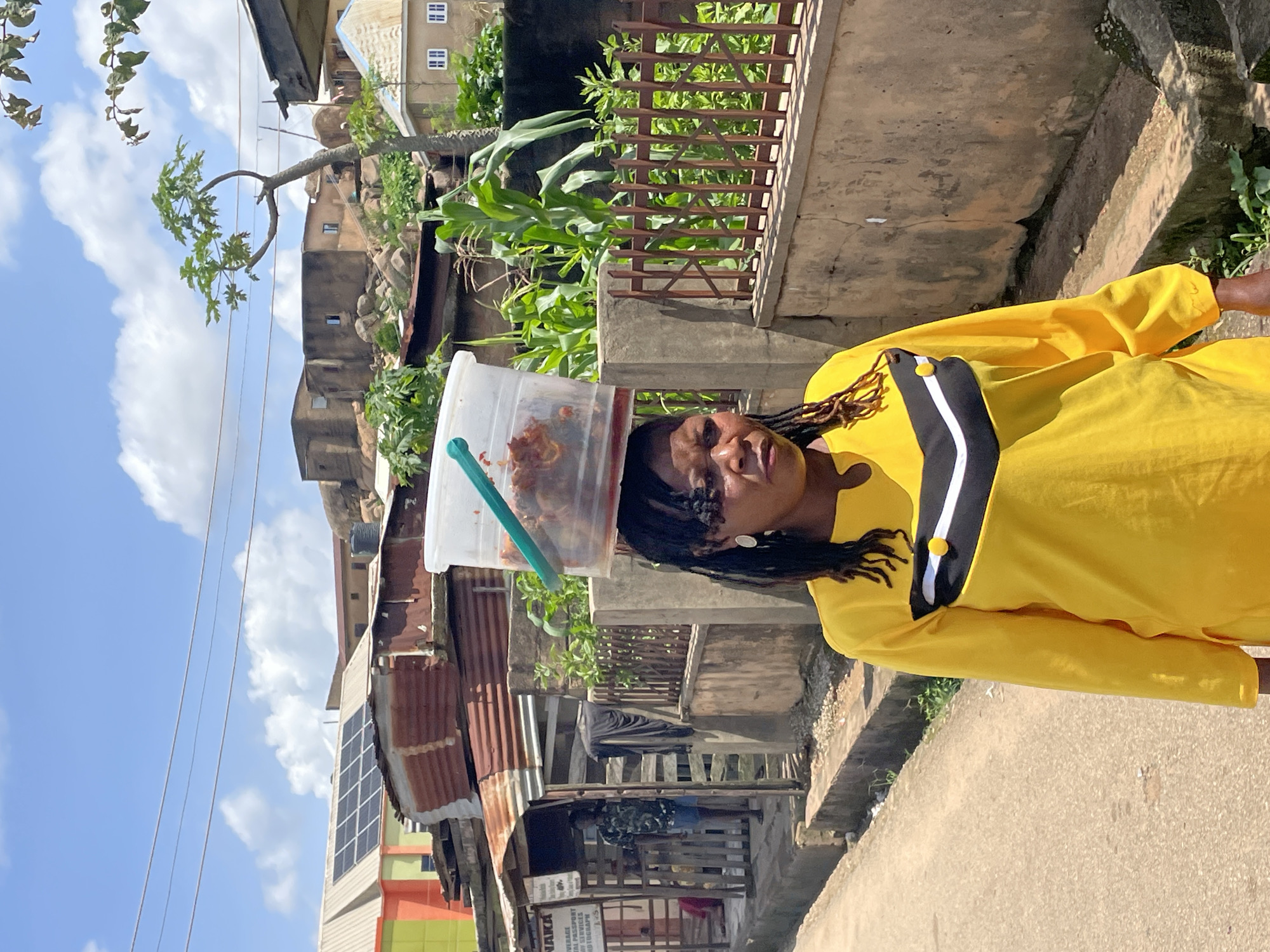 A woman in a bright yellow tunic walks down a dirt path in Jos, Nigeria, carrying a clear plastic bucket filled with food on her head. The bucket has a green handle and contains what appears to be cooked meat or stew. The woman has dark skin, braided hair, and wears large silver earrings. She is looking forward with a neutral expression. Behind her, a weathered wooden fence encloses a yard with green corn stalks. In the background, several buildings with corrugated metal roofs and concrete walls are visible, some built into rocky hillsides. The sky is blue with scattered white clouds, and power lines stretch across the frame. The scene suggests a typical day in a residential area of Jos.