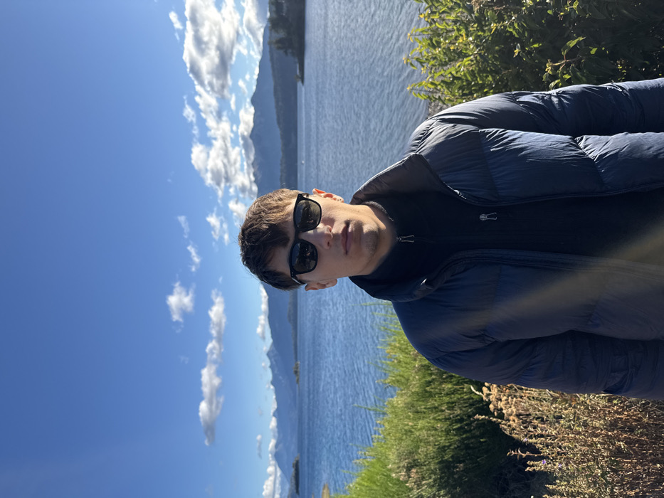 A man stands outdoors wearing sunglasses and a navy puffer jacket. Behind him is a body of water and mountains under a blue sky with scattered clouds. Green shrubbery is visible in the foreground. The man is facing slightly off-center. The photo appears to be taken during the day with clear lighting.